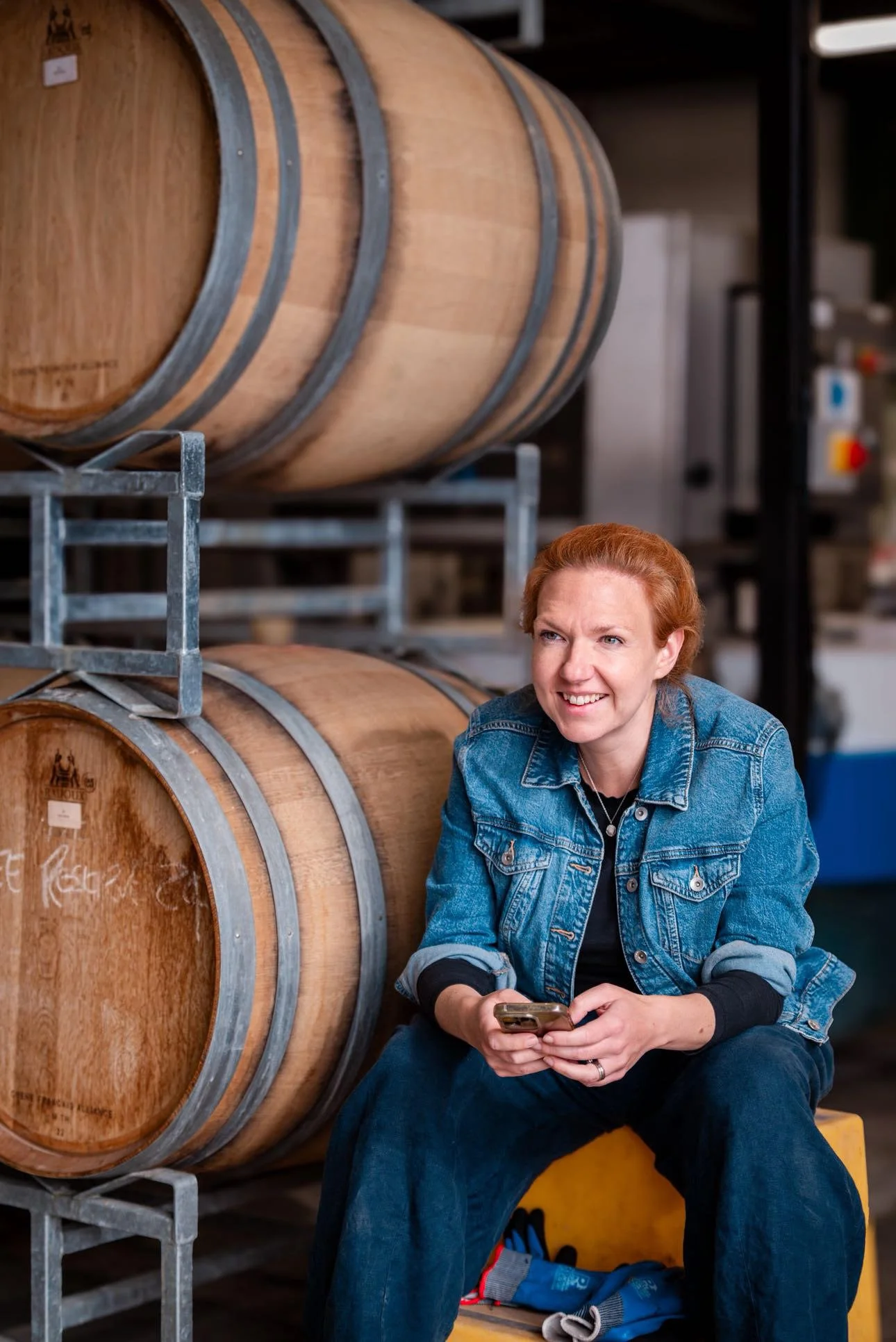 A woman with red hair wearing a denim jacket sitting on a yellow box, holding a smartphone, in front of wine barrels in a winery or cellar.