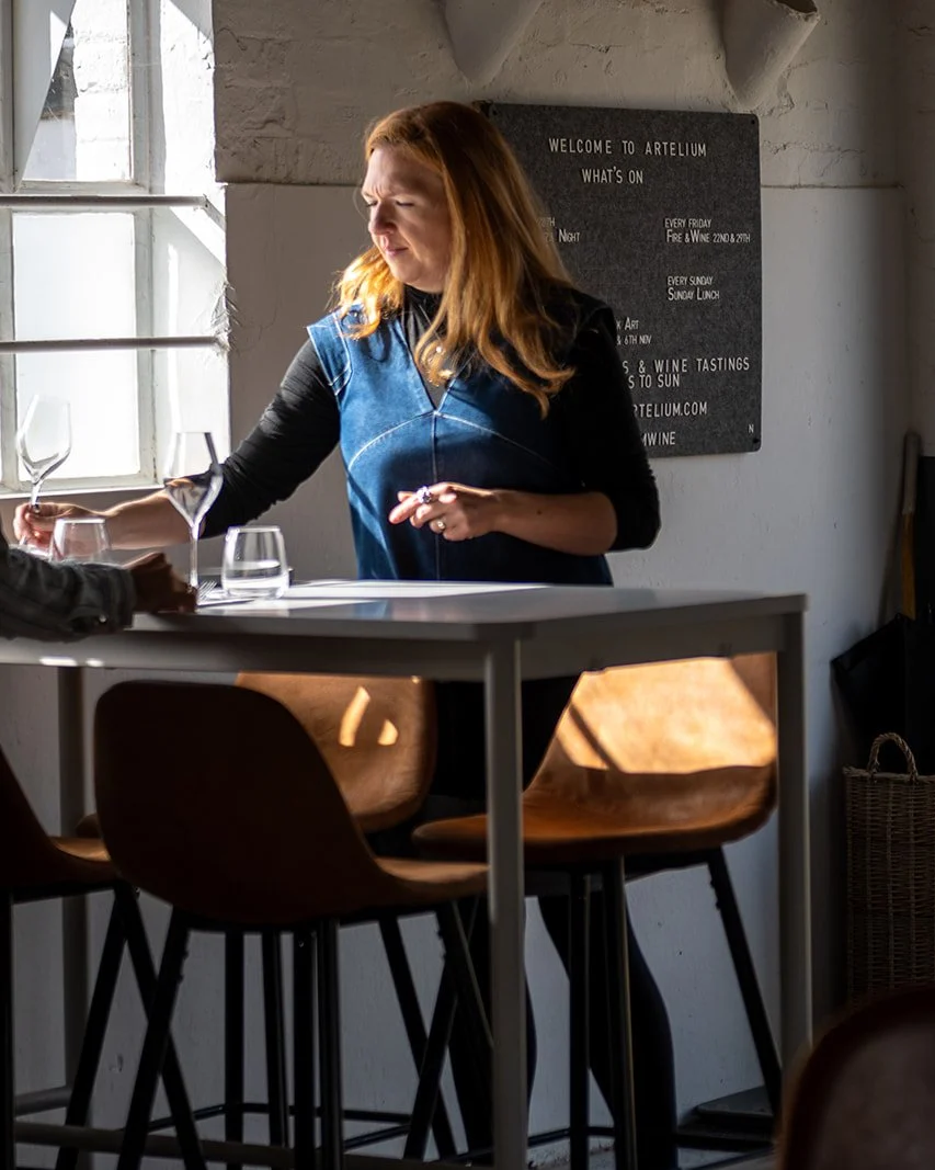 A woman with red hair wearing a blue apron is setting wine glasses on a table in a winery or tasting room. Sunlight streams through a window, casting shadows on the table and chairs. A blackboard in the background lists events and tastings at Arteliu.