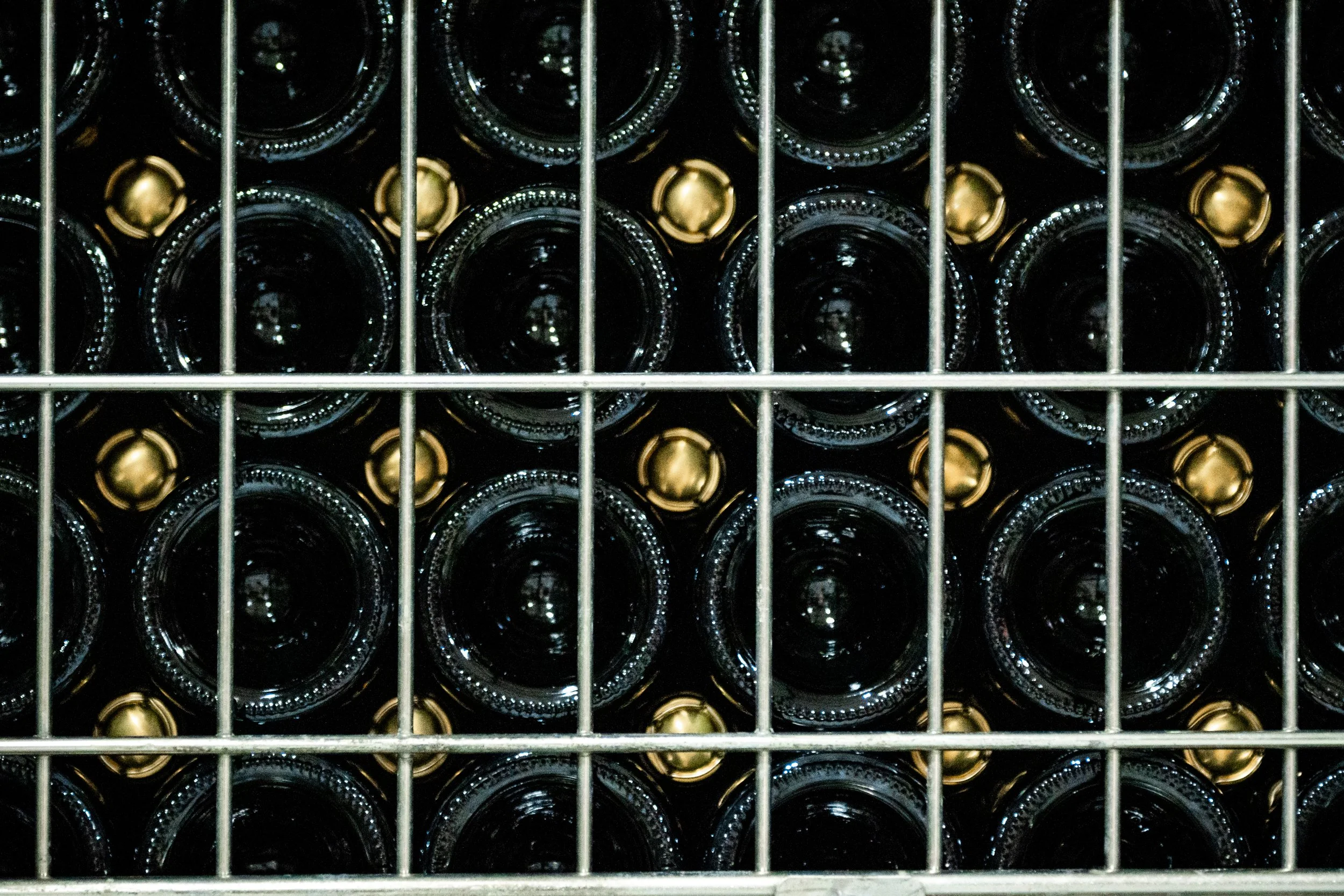 A top view of wine bottles stacked horizontally in a bottle rack, showing the necks with gold-colored caps through the metal grid.