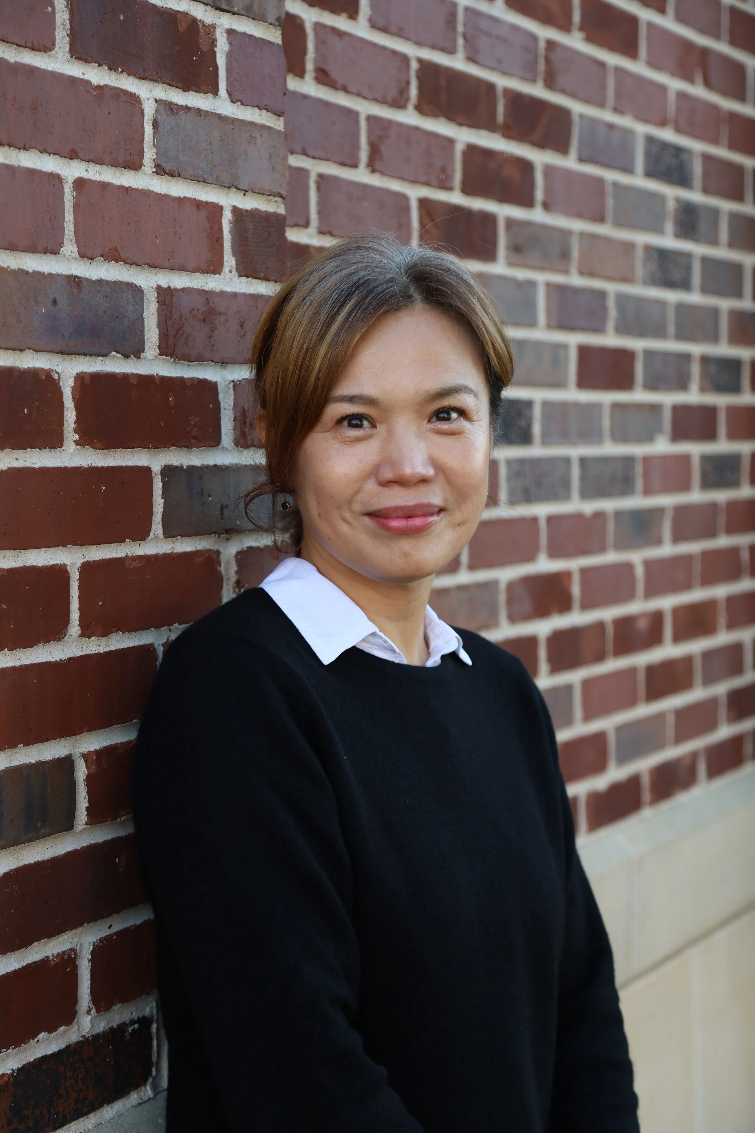 Woman with short brown hair smiling, wearing a white shirt and black sweater, standing beside a brick wall.