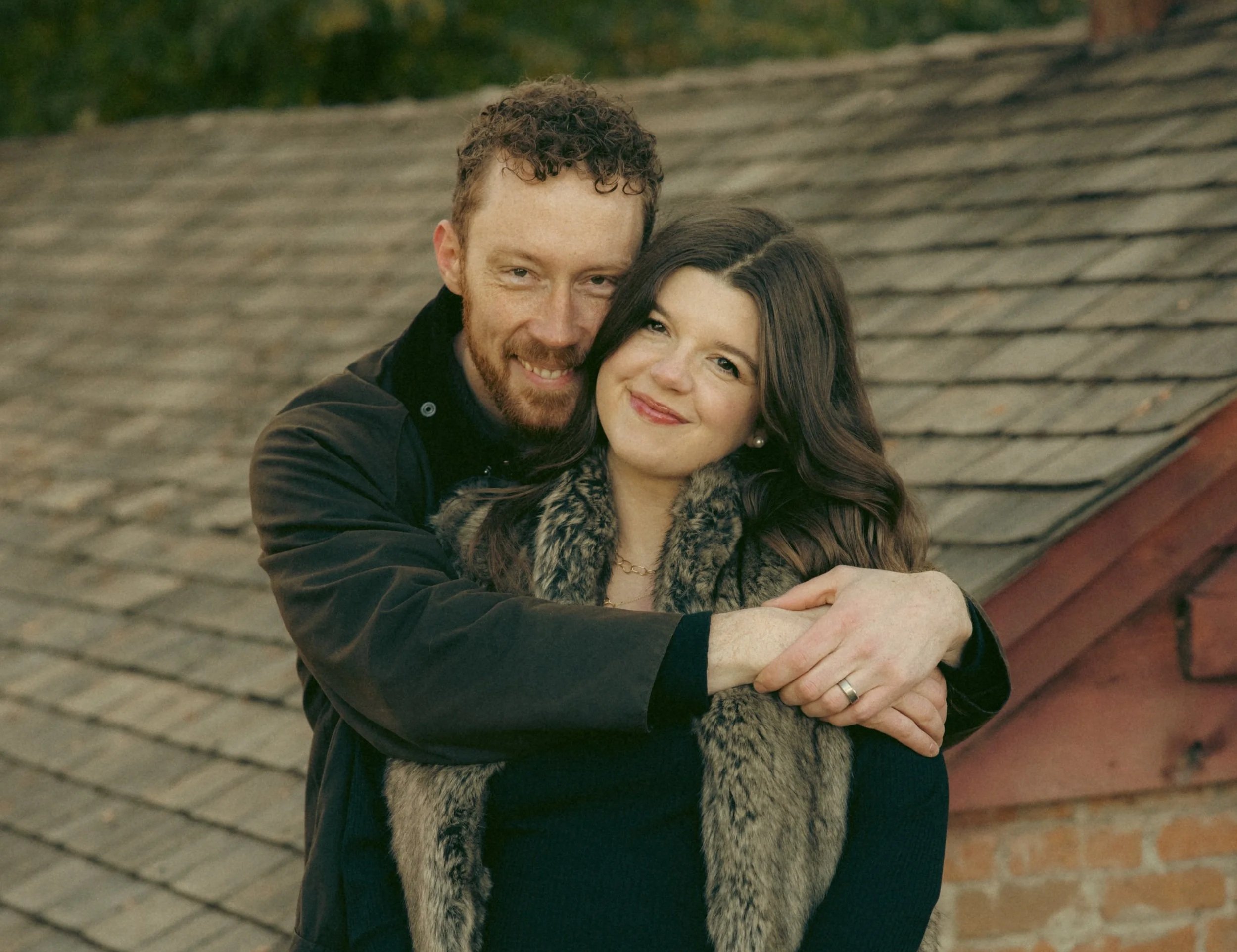 A couple hugging outdoors on a brick patio, with a blurred background of trees and a roof.