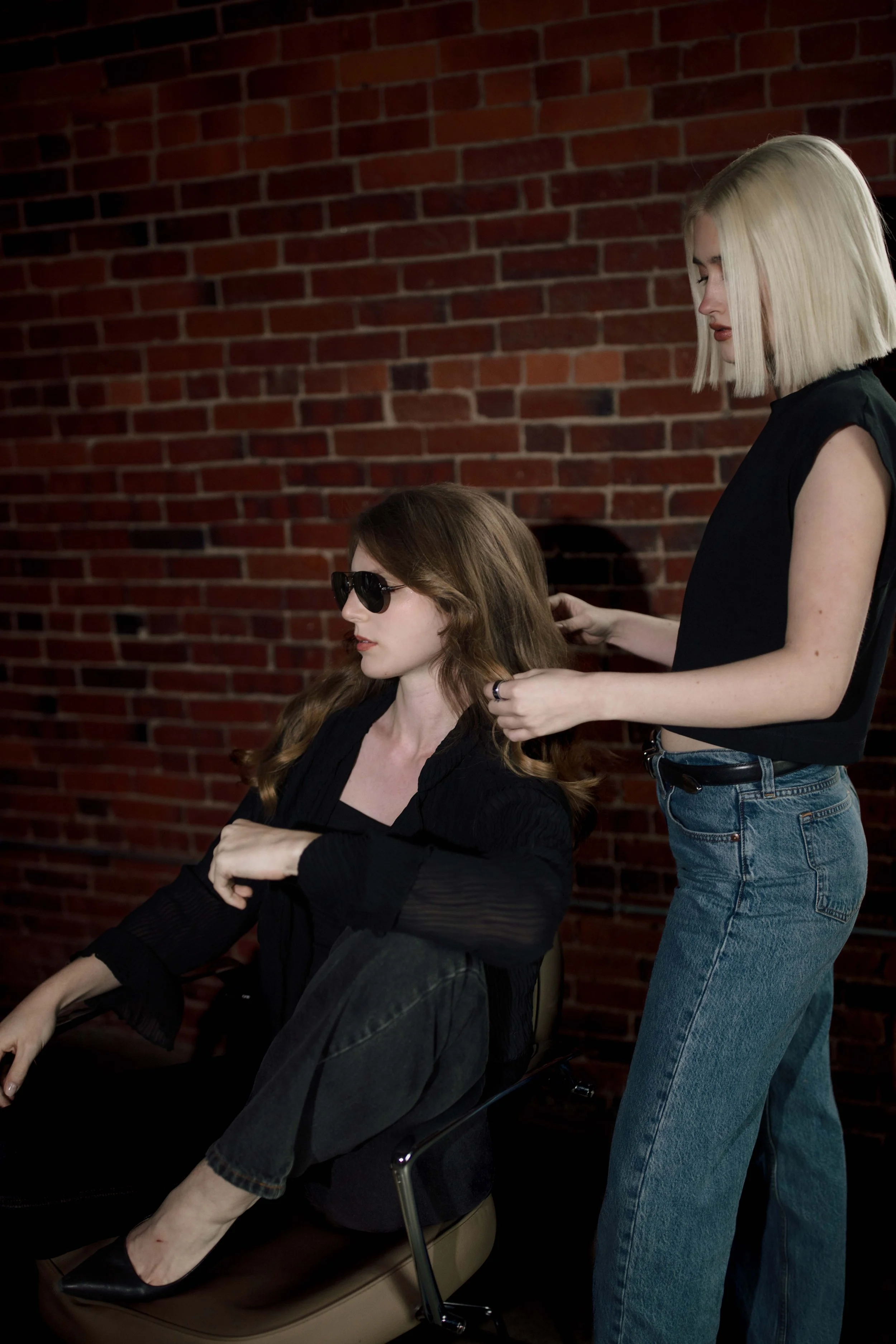 A woman with long brown hair and sunglasses sitting in a chair with crossed arms while another woman with blond hair styles her hair, in front of a red brick wall.