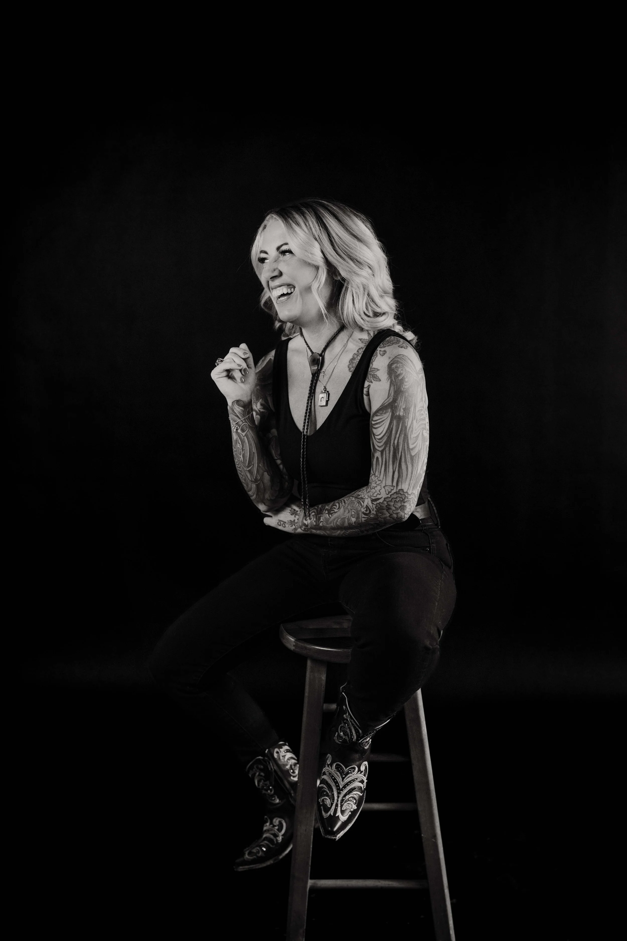 A woman with blonde hair and tattoos on her arms laughing while sitting on a stool against a black background.