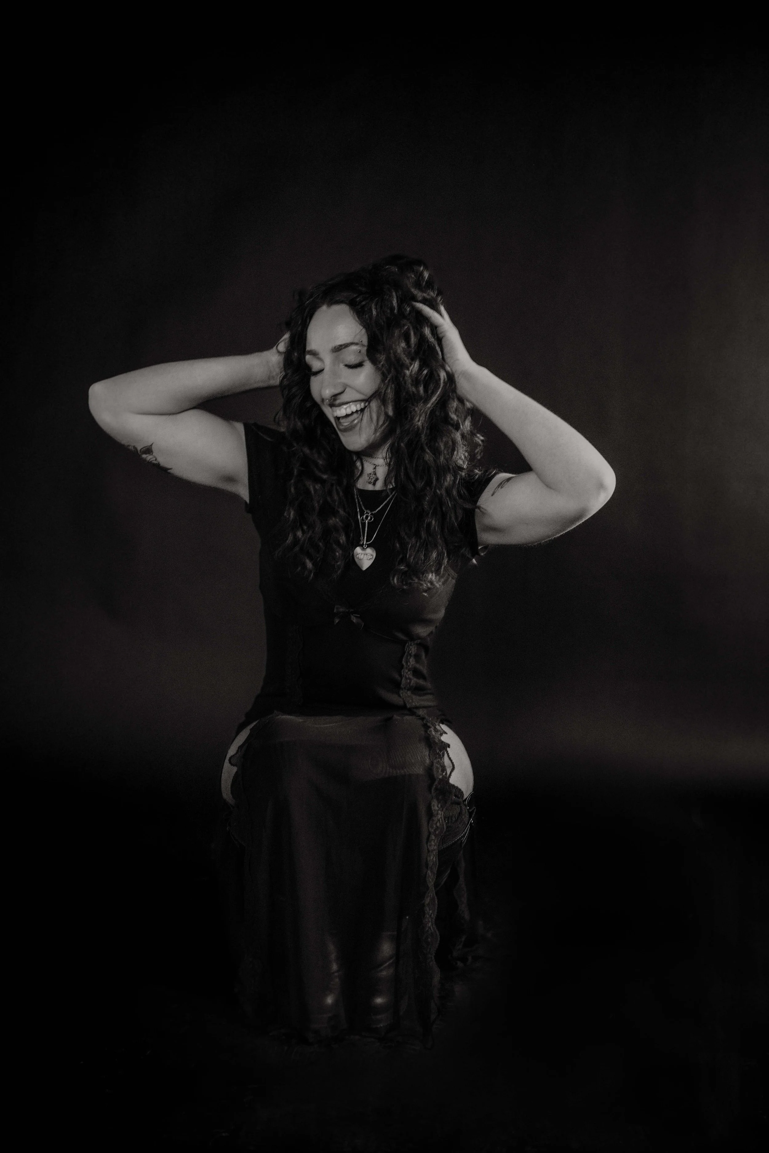 A woman with curly hair, smiling and laughing, wearing a black top and layered necklaces, holding her head with both hands, against a dark background.