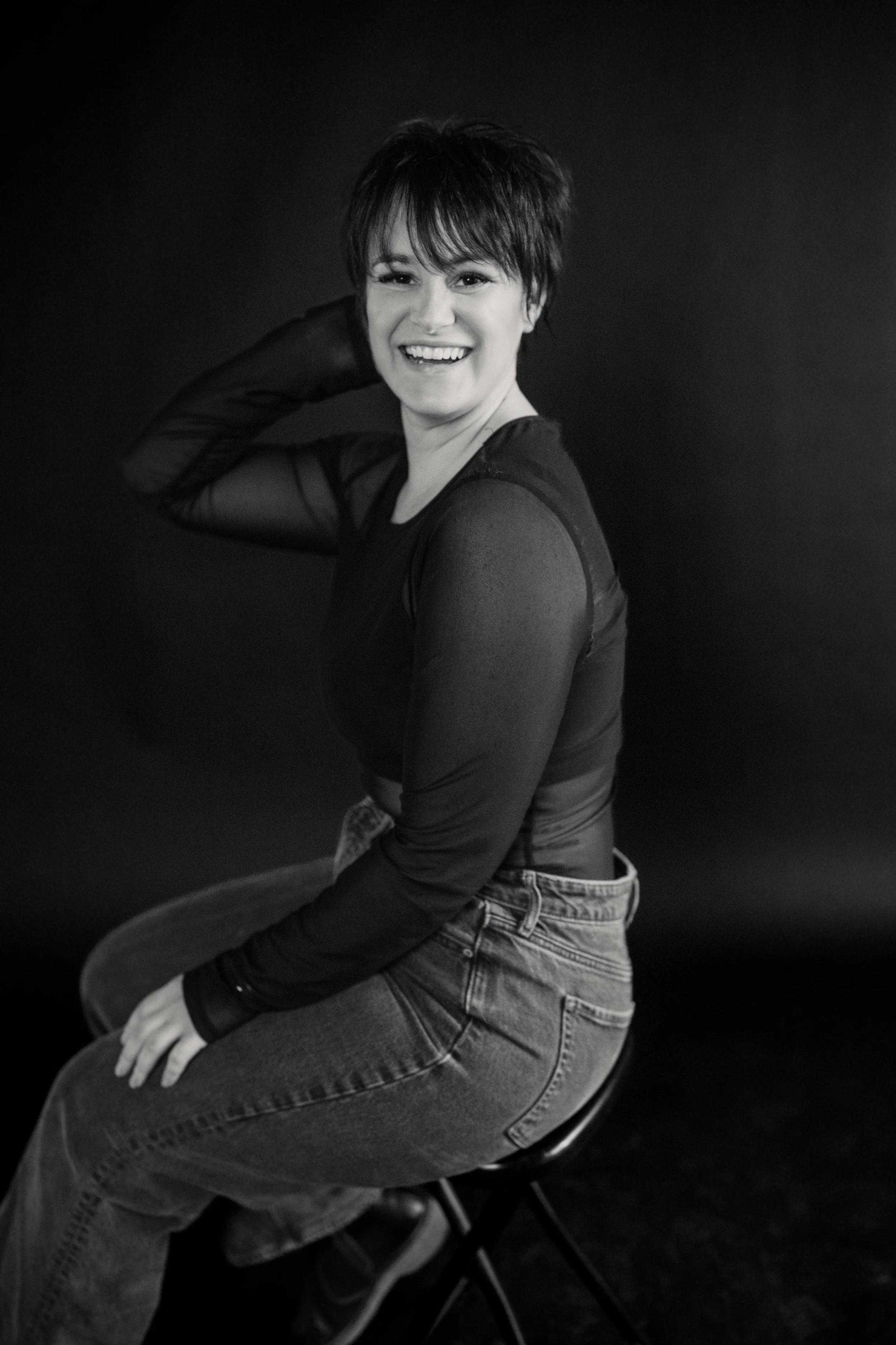 A woman with short dark hair smiling and sitting on a black chair, wearing a long-sleeved black top and jeans, against a black background.