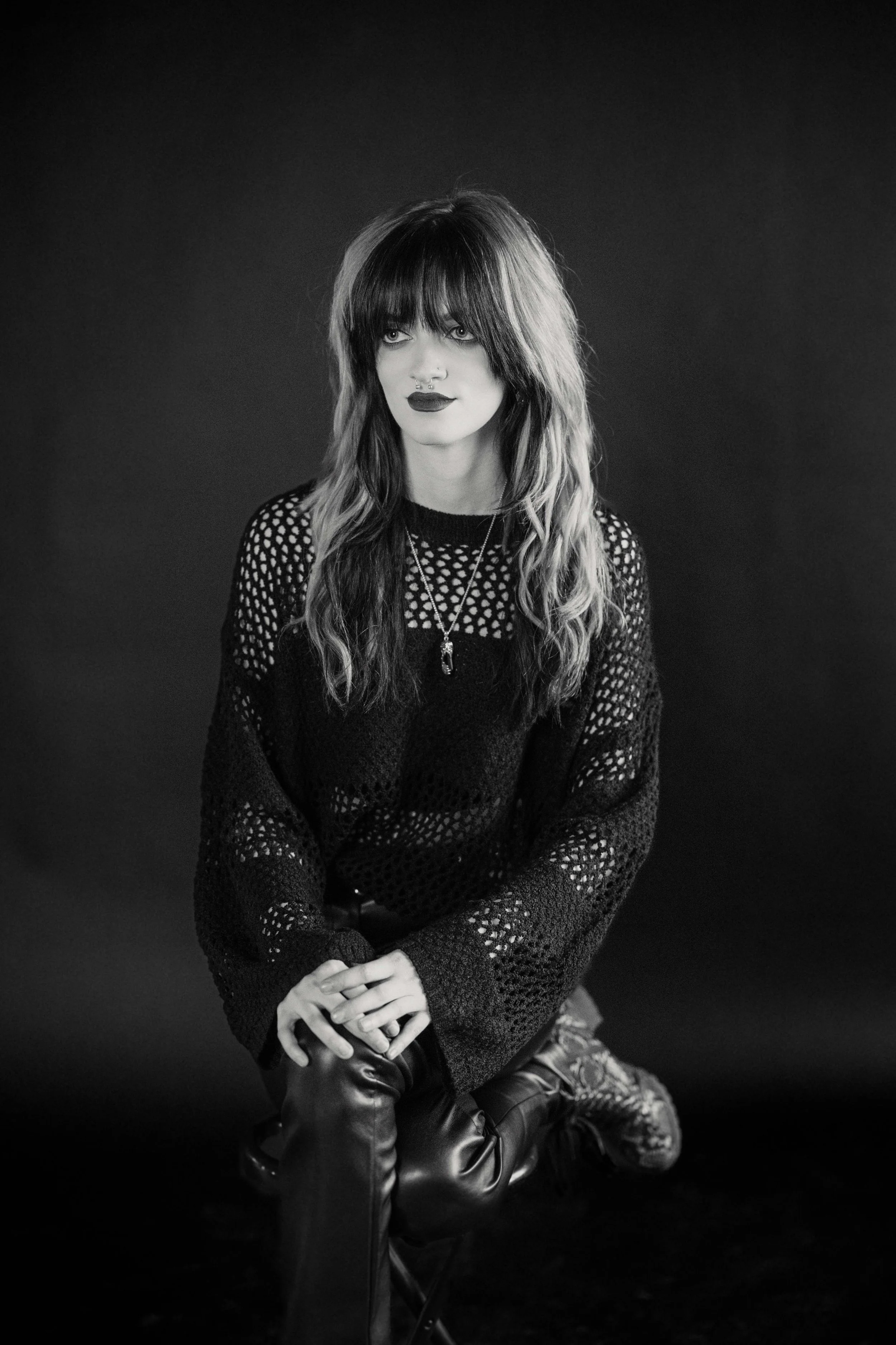 A young woman with long wavy hair, bangs, and dark lipstick, wearing a black, open-knit sweater and layered necklaces, sitting on a stool with her hands clasped, against a plain dark background.