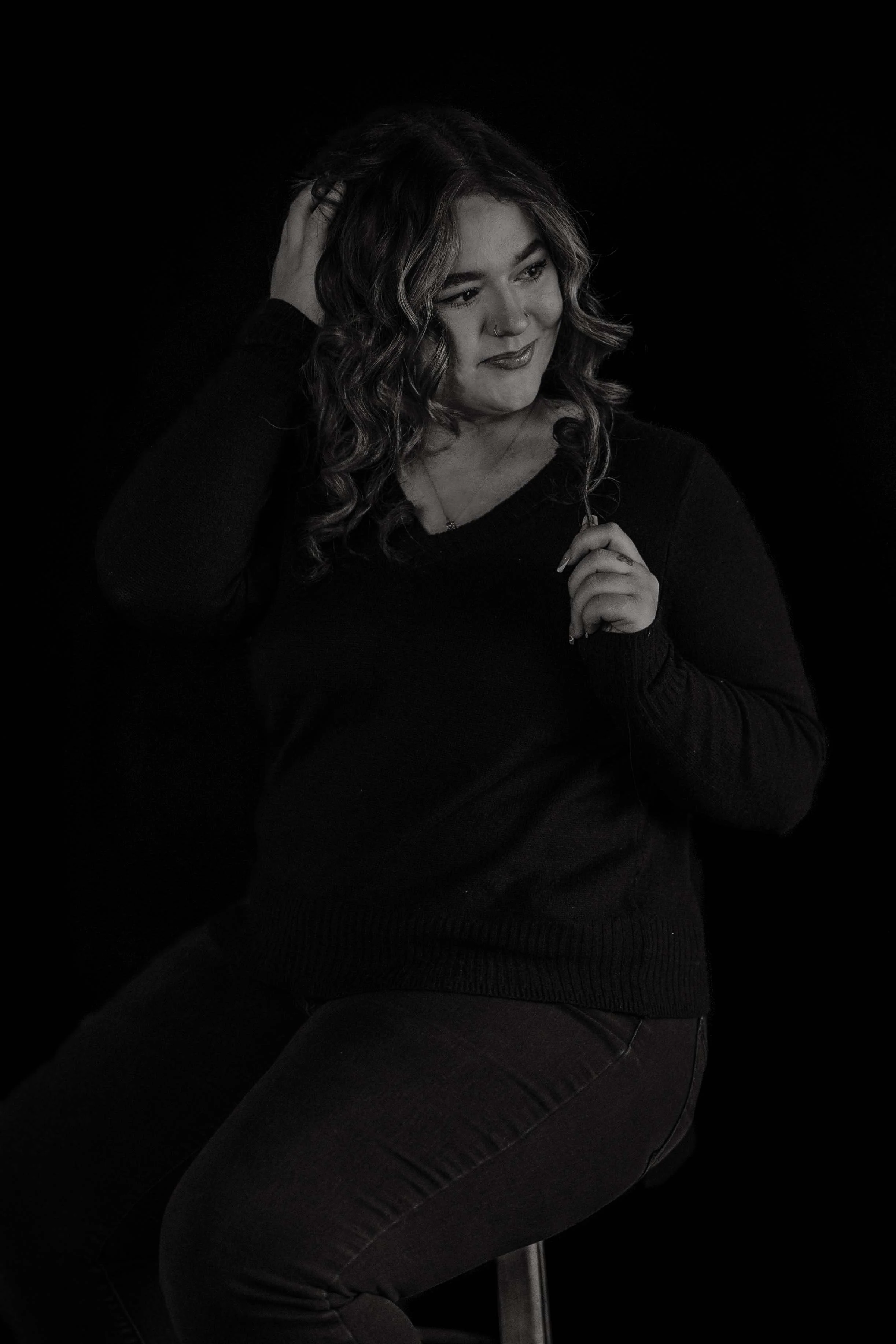 A woman with curly hair, wearing a black long-sleeve top, sitting against a dark background, touching her hair with her left hand and holding a small necklace with her right hand, smiling softly.