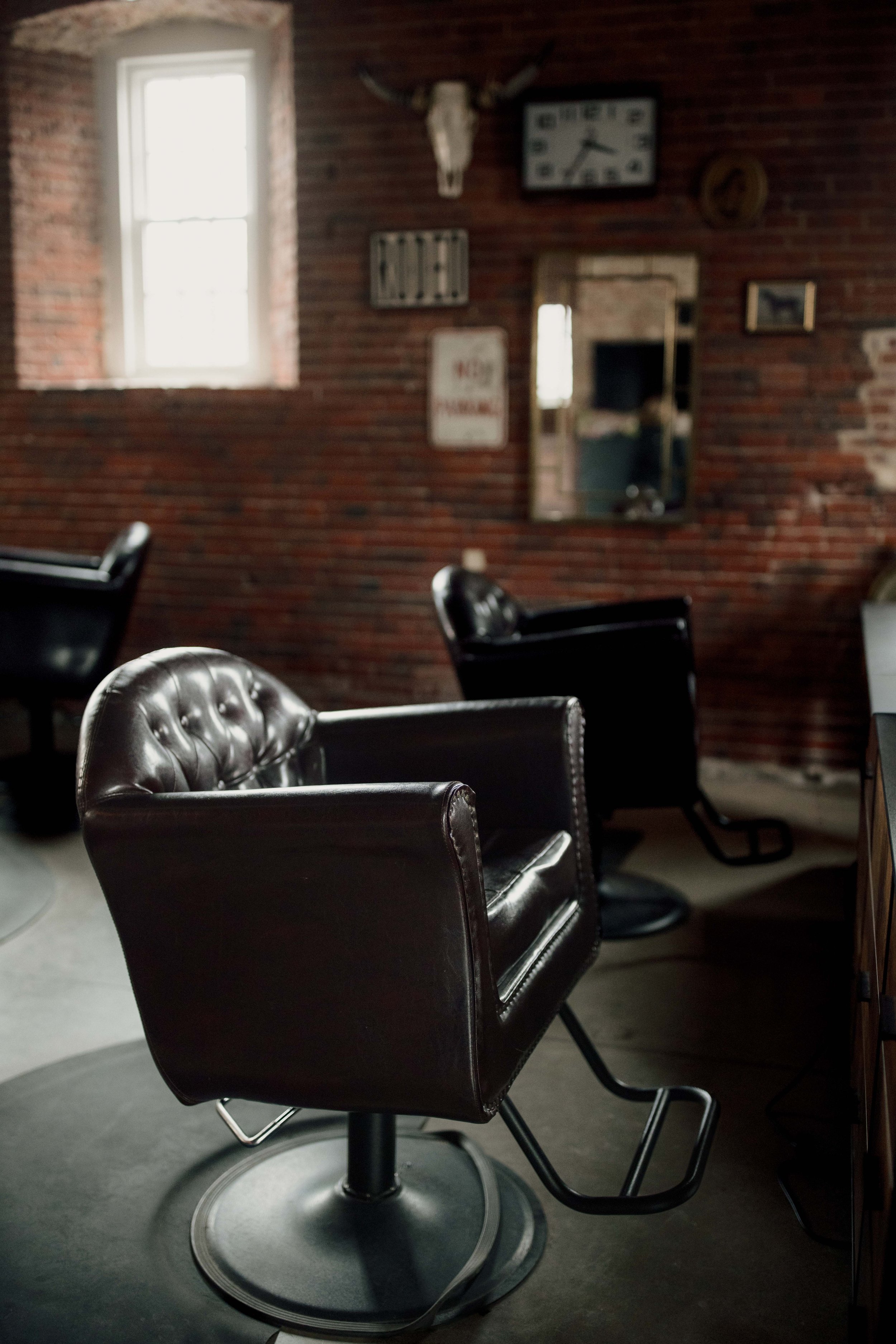 A room with brick walls and natural light from a window. There are black leather chairs with metal armrests and bases, and vintage decor including a skull of a cow, a clock, and various framed pictures.