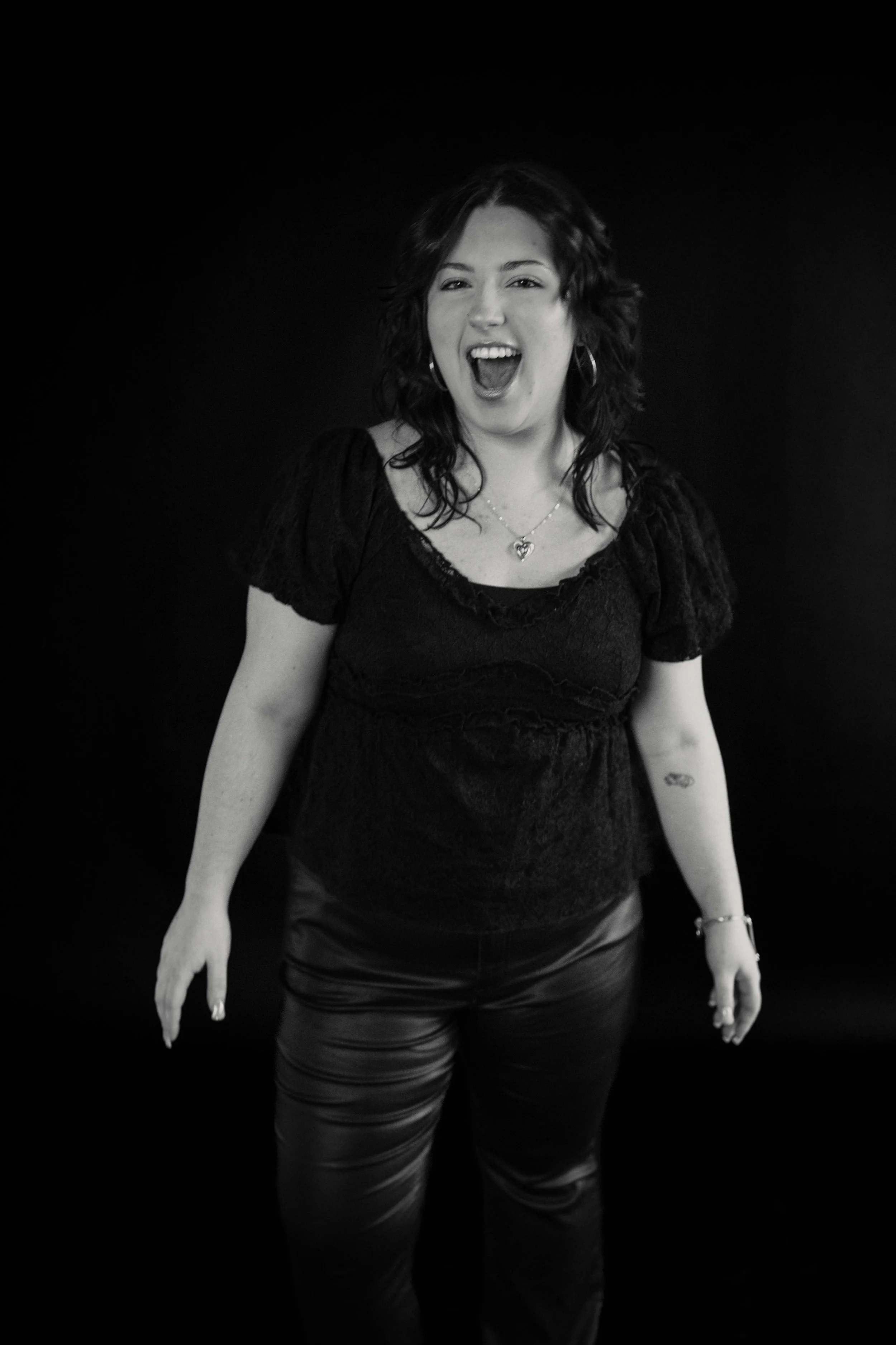 A woman with wavy dark hair wearing hoop earrings, a necklace, a lace black top, and leather pants, standing against a plain dark background, smiling and looking cheerful.