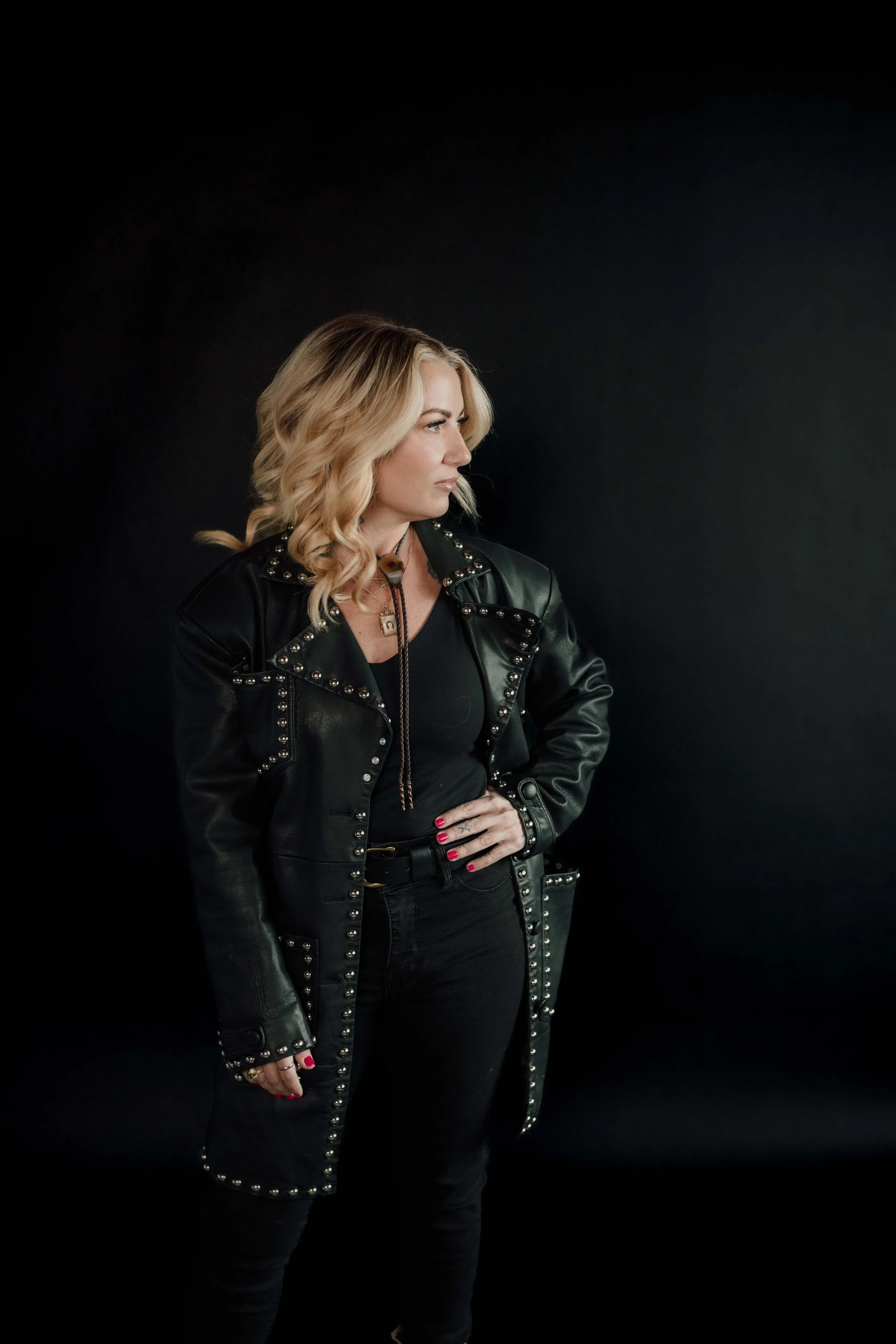 A woman with blonde wavy hair wearing a black leather jacket with silver studs, black top, and black pants, standing against a black background, looking to her left with her hand on her hip.