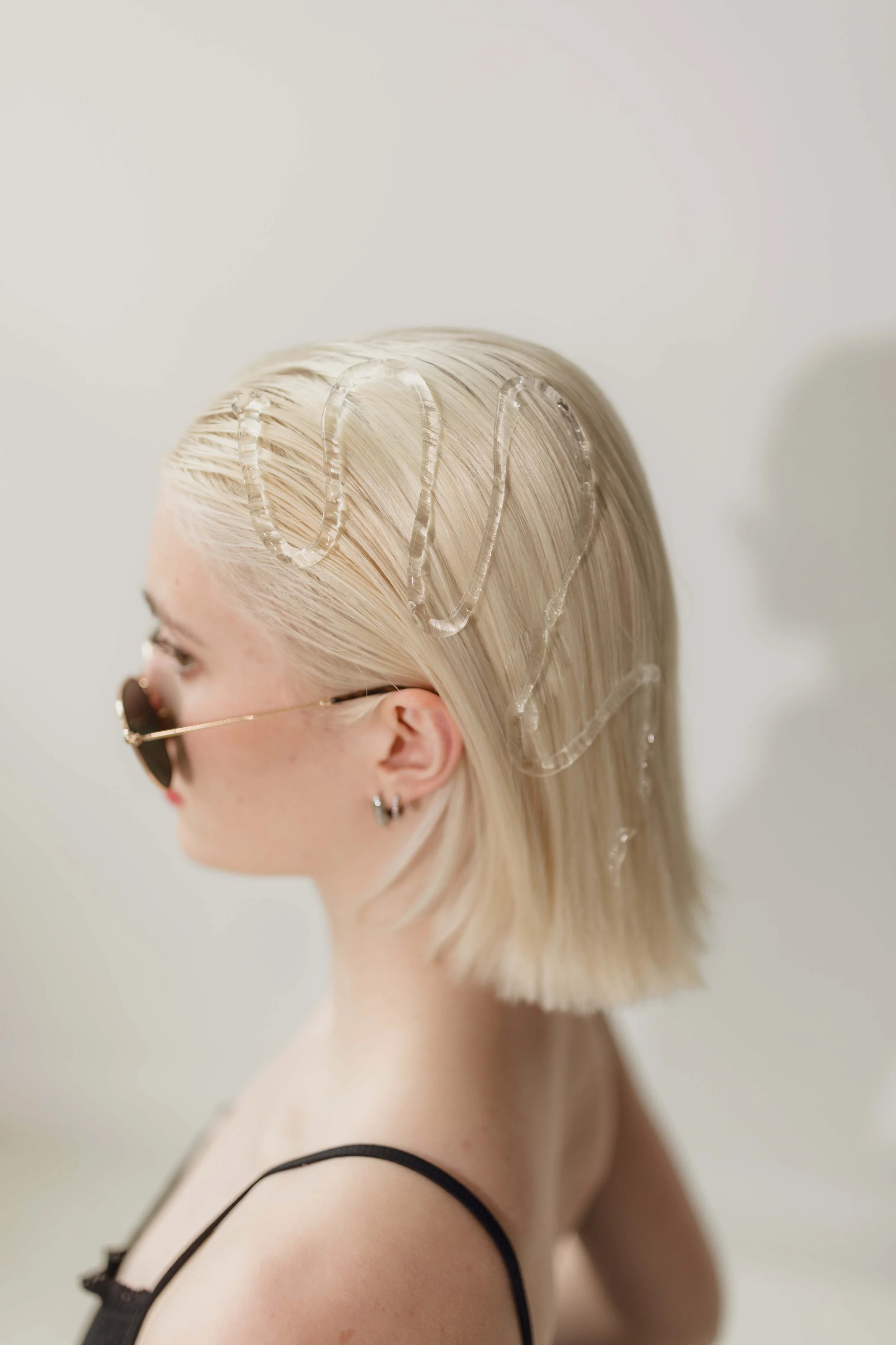 A woman with platinum blonde hair wearing sunglasses, a black spaghetti strap top, and earrings, looking to the side against a plain background with a shadow.