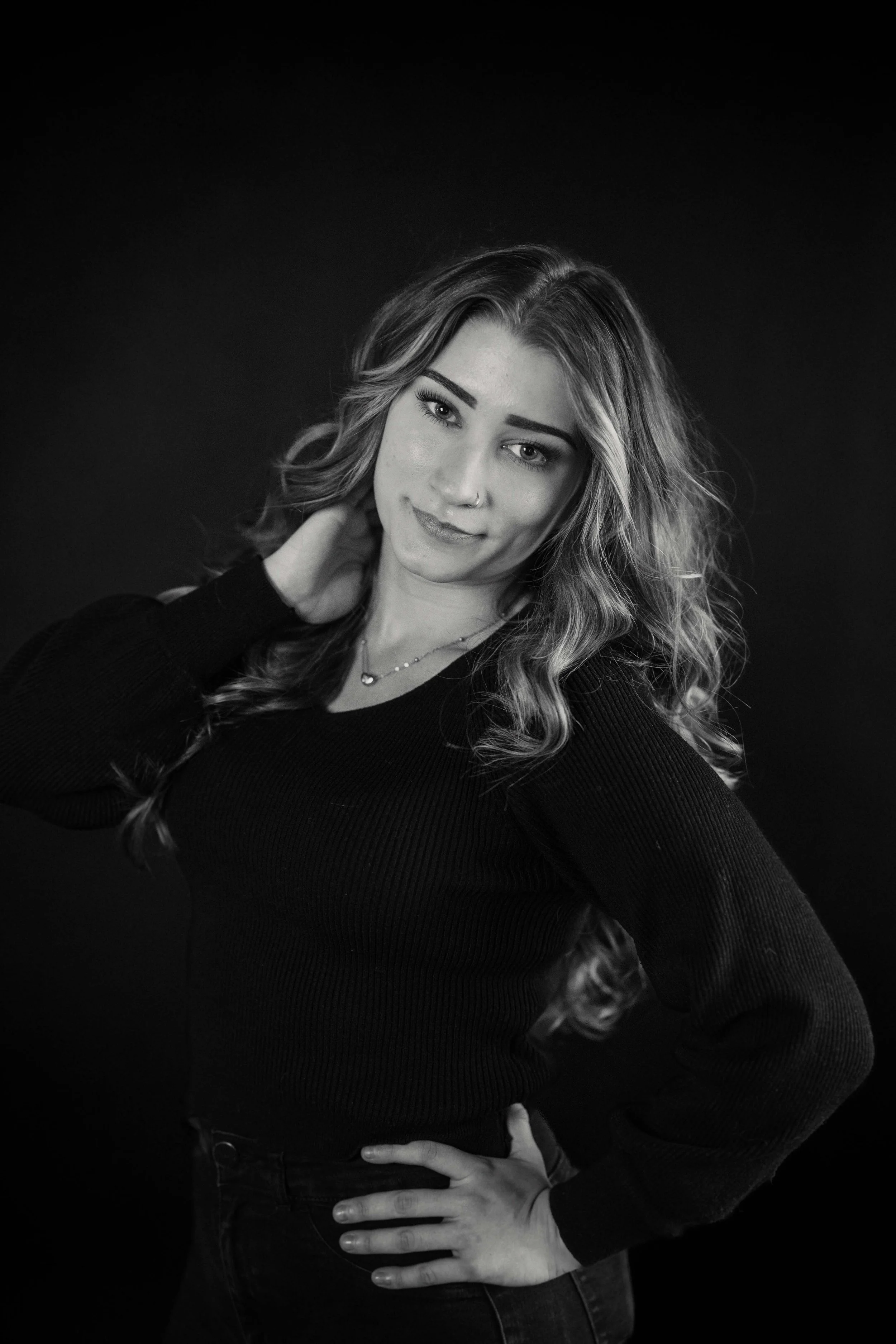 Black and white photo of a young woman with wavy hair, wearing a long-sleeve black top, posing with one hand on her shoulder and the other on her hip.