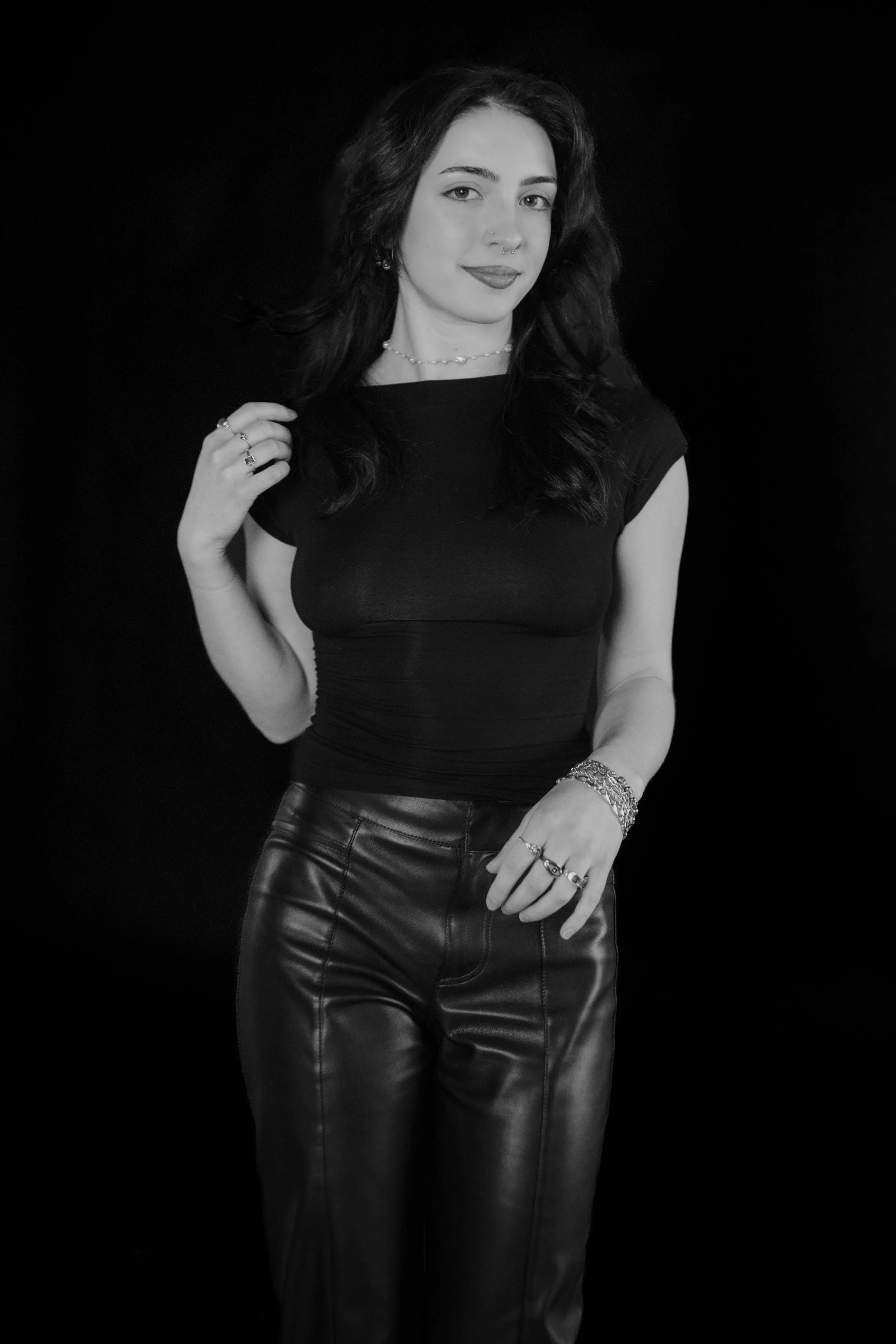 Black and white portrait of a young woman with dark hair, wearing a black top and leather pants, adorned with jewelry including rings, necklaces, and bracelets, standing against a plain dark background.