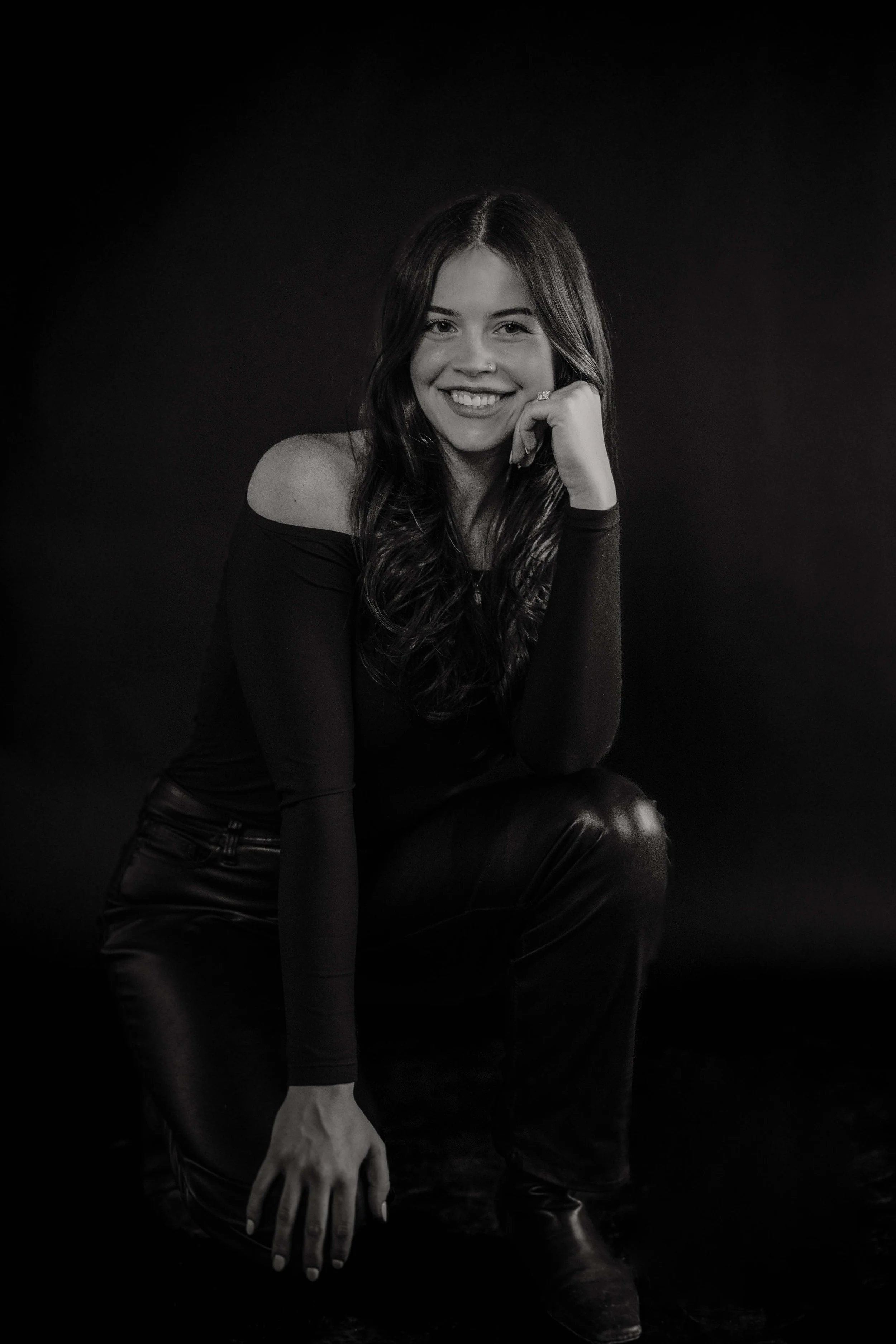 A black-and-white photo of a woman smiling, sitting with one hand on her knee and the other hand resting on her cheek. She has long, wavy hair, and is wearing a long-sleeved top and leather pants. The background is dark.