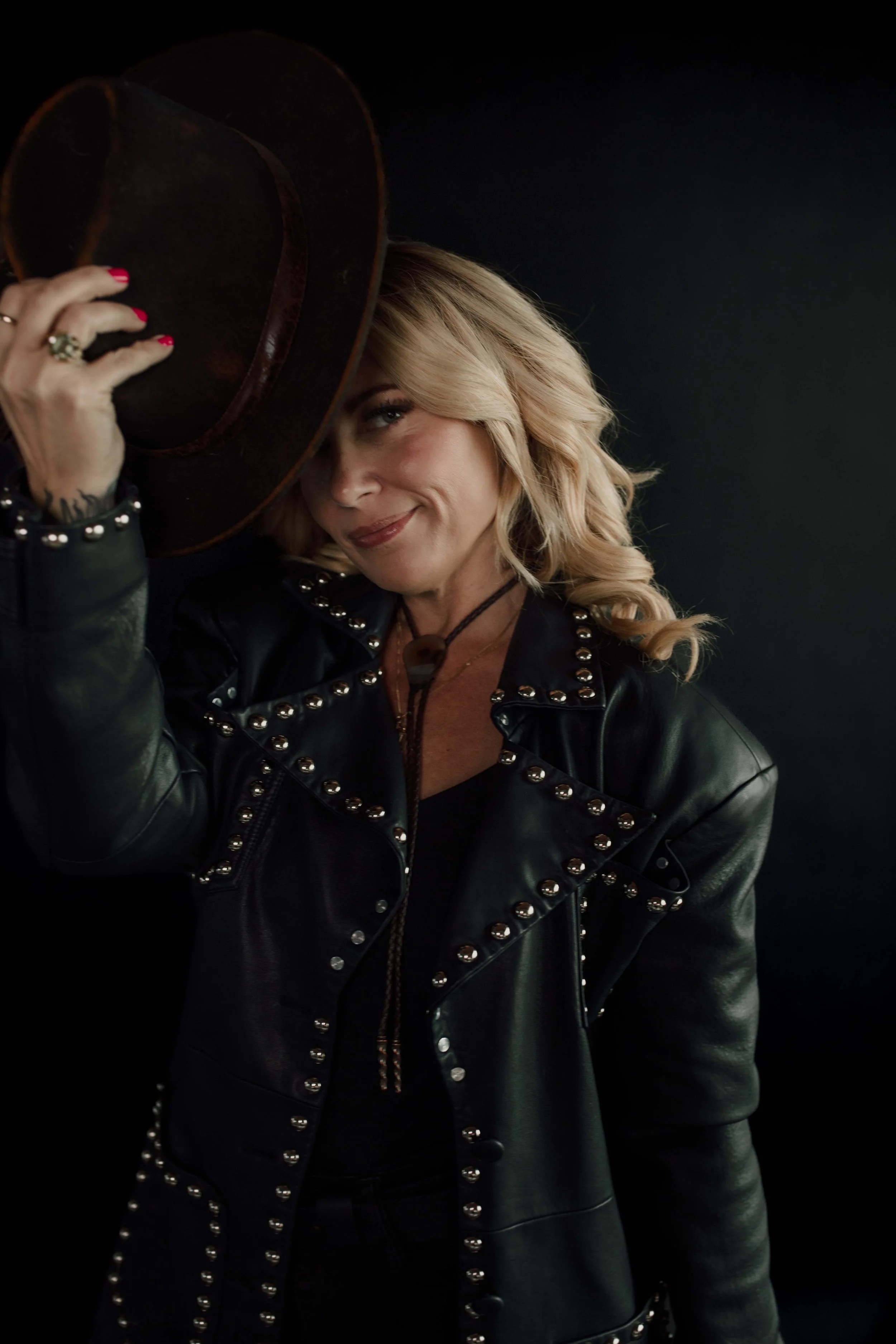 A woman with blonde hair and a leather jacket with silver studs, holding a hat to her head and smiling against a black background.