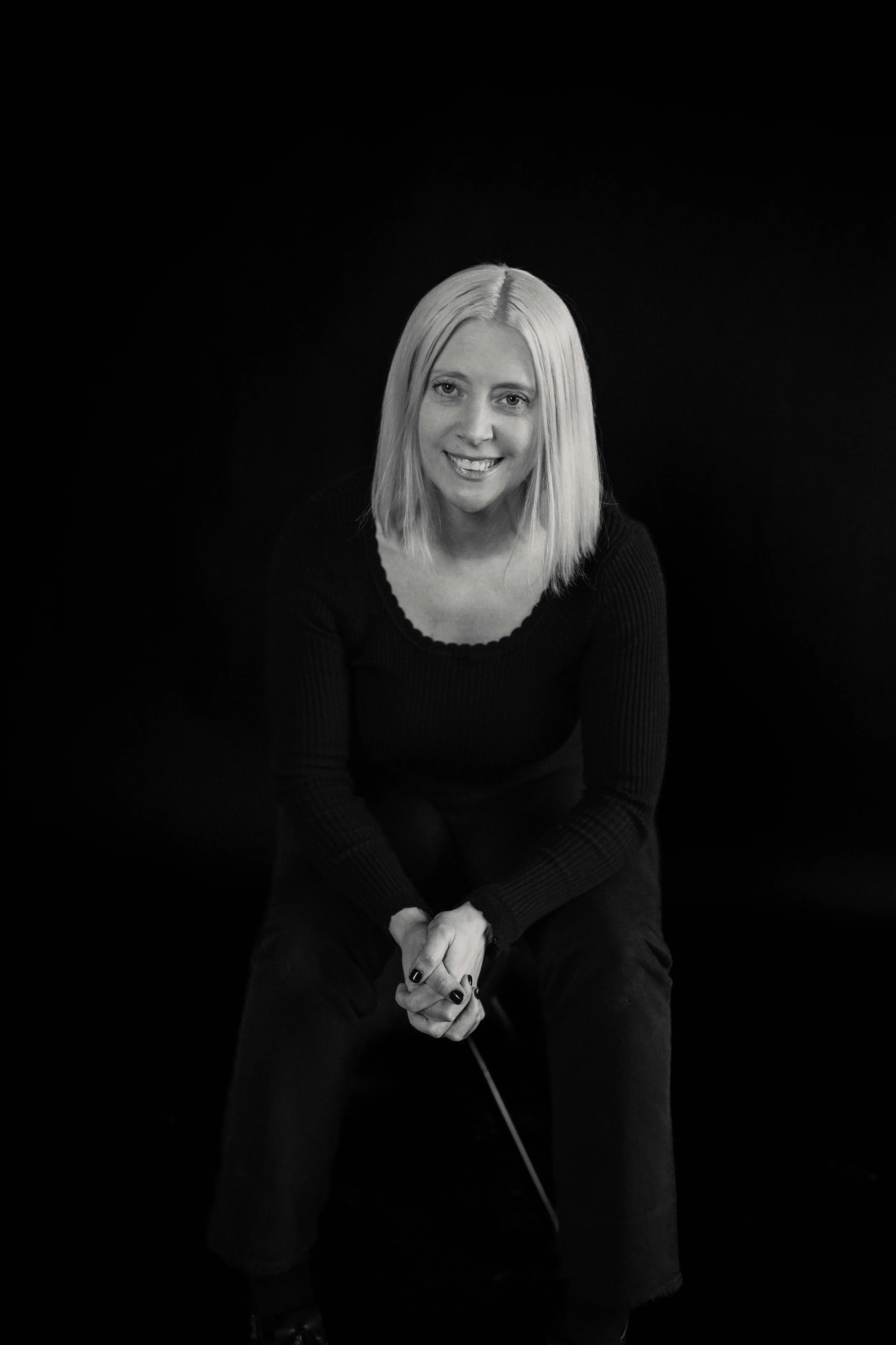 A woman with shoulder-length blonde hair, sitting against a black background, smiling at the camera with her hands clasped in her lap.