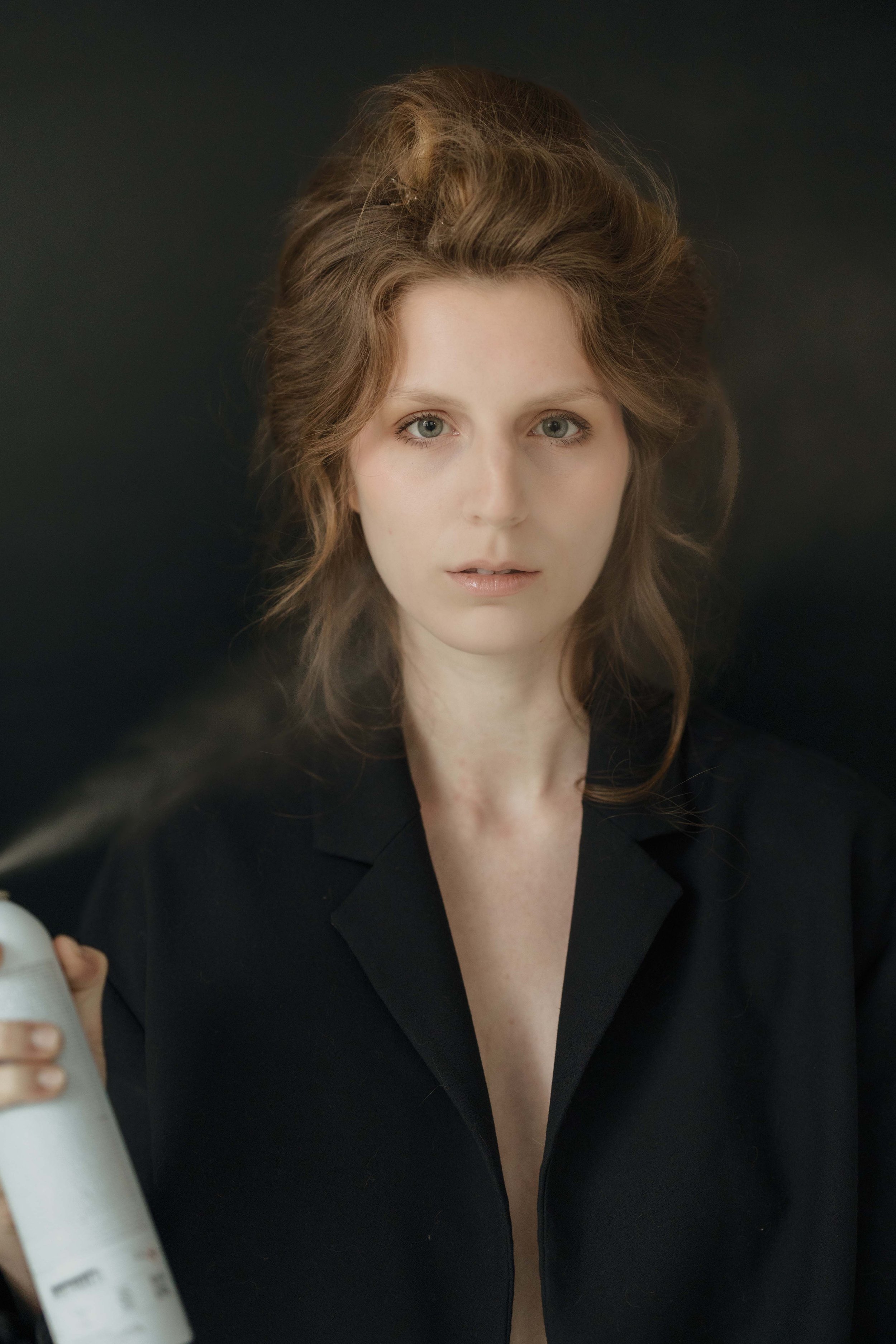 A woman with light skin and curly, brown hair wearing a black blazer with a deep neckline, looking directly at the camera against a black background. She holds a white spray bottle with a spray nozzle in her left hand, which is partially visible in the foreground.