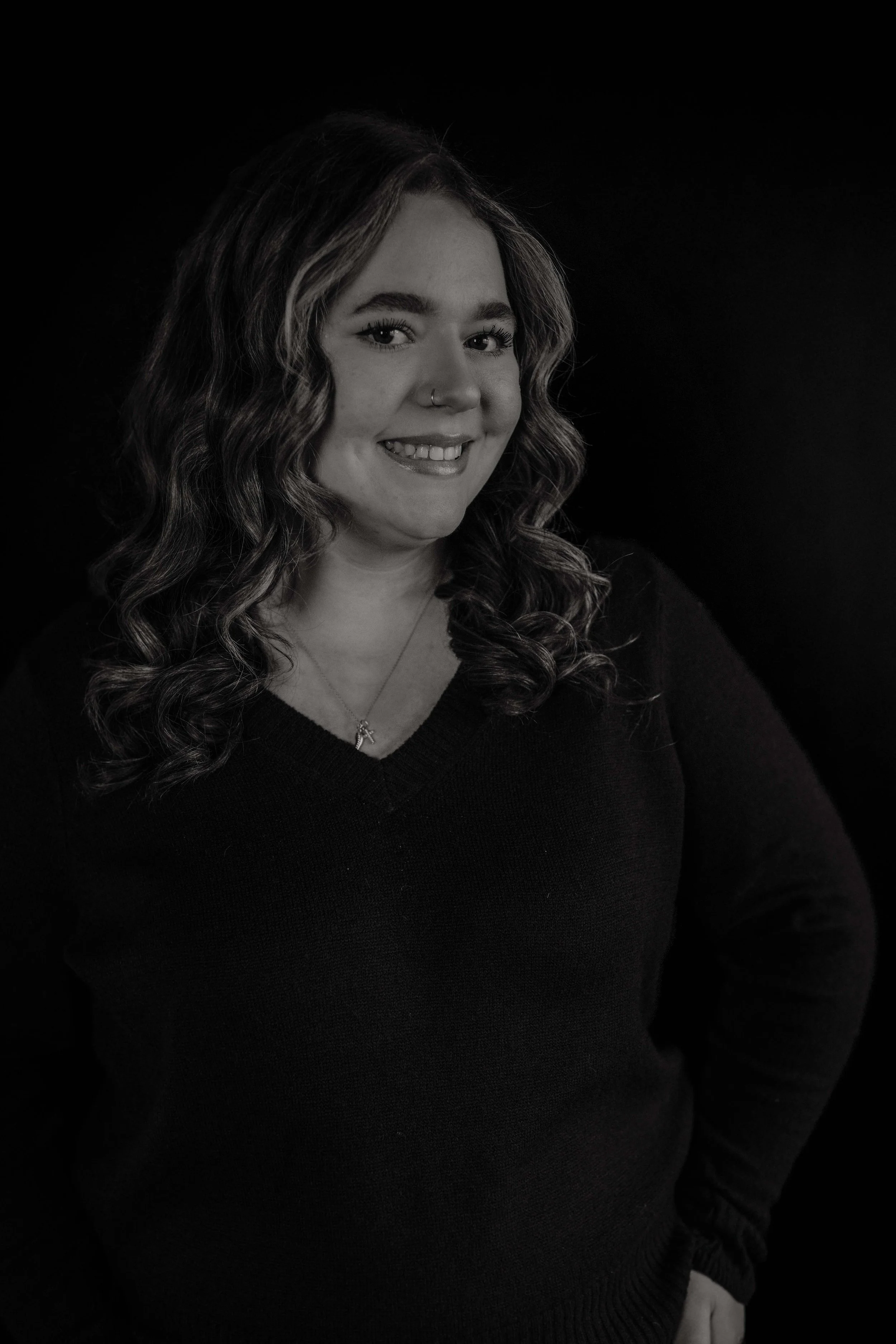 Black and white portrait of a woman with long wavy hair, smiling, wearing a necklace and a dark sweater, against a dark background.