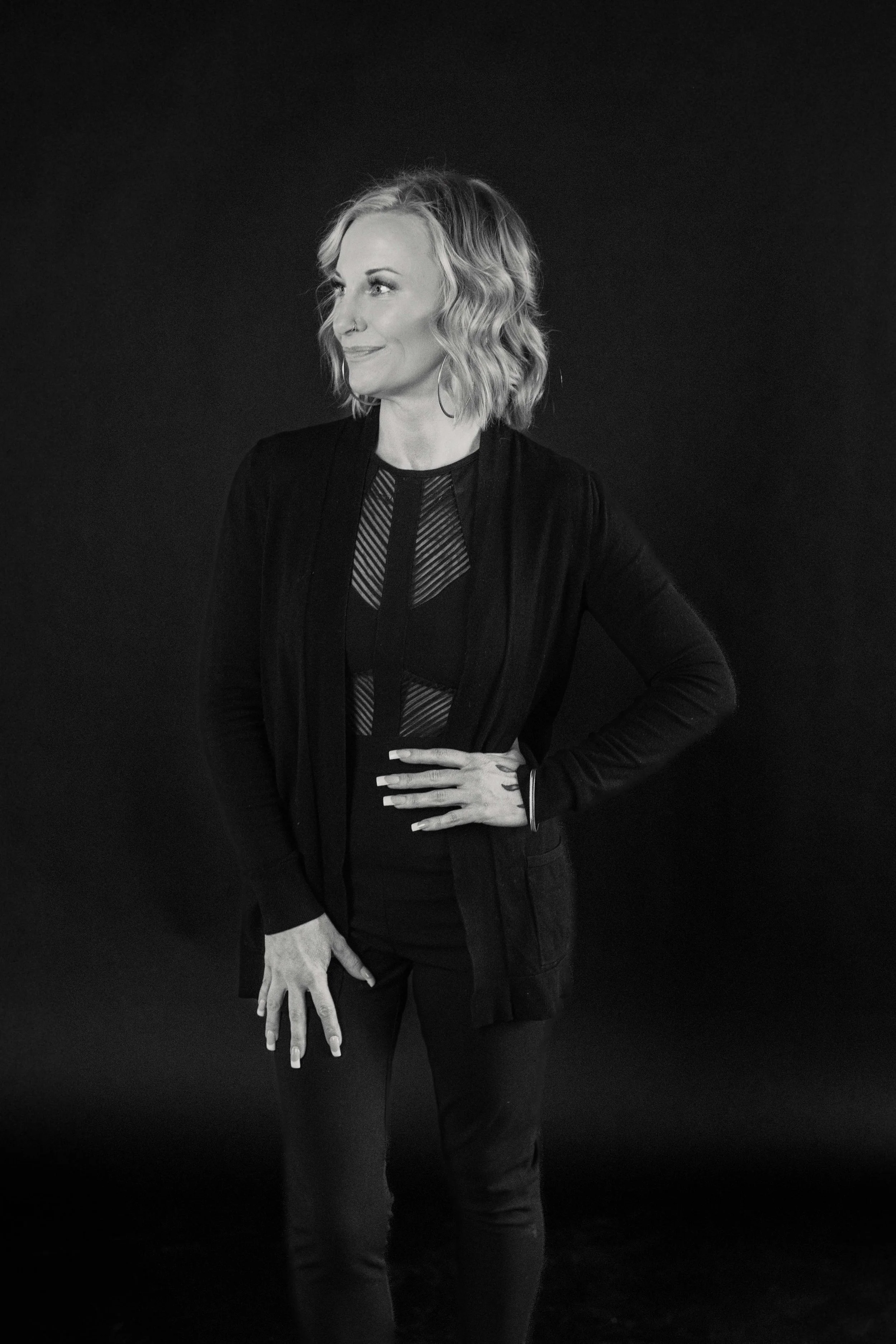 Black and white photo of a woman with wavy hair, wearing a dark blazer over a patterned top, standing against a dark background, with her left hand on her hip and her right hand resting on her thigh.