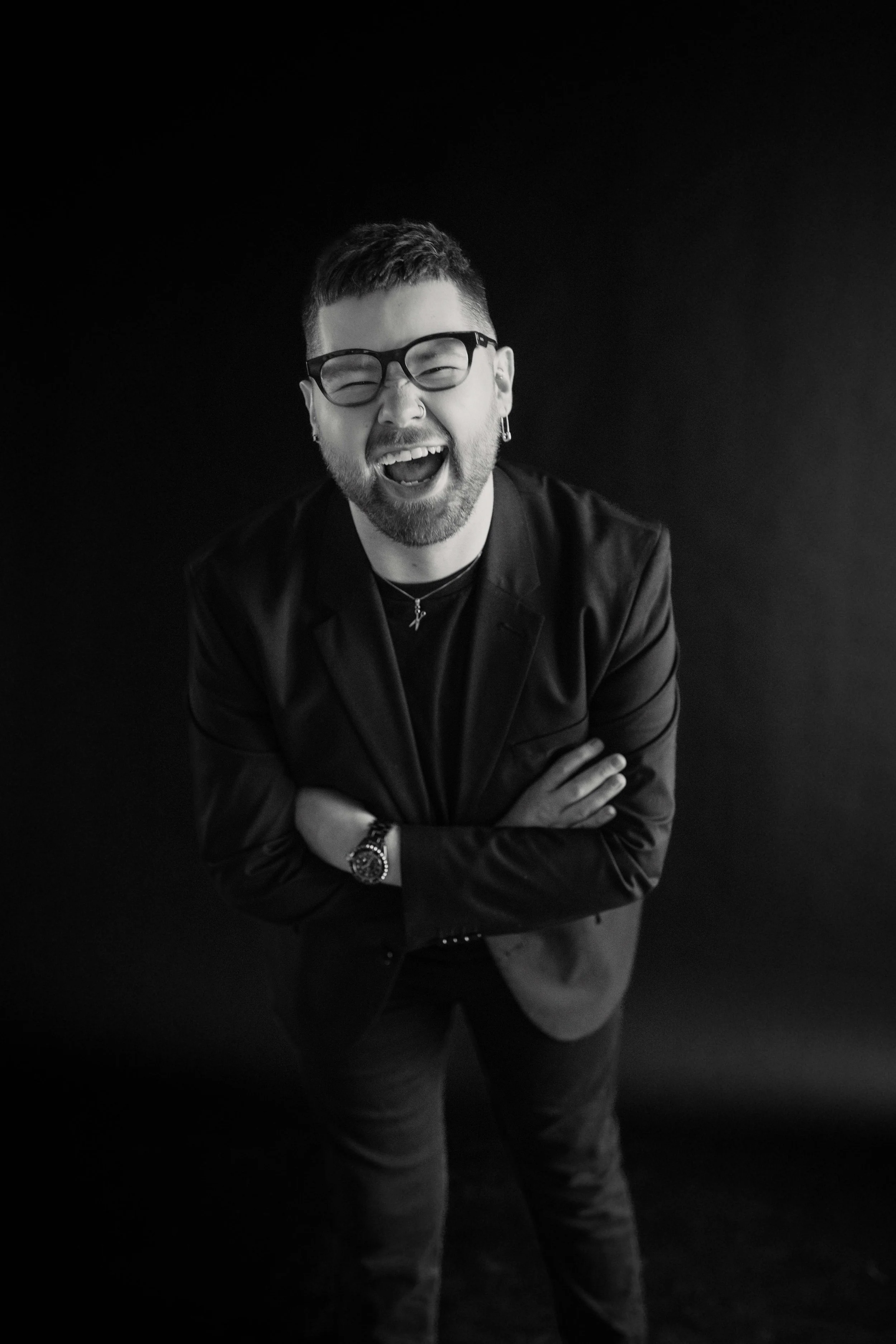 Black and white photo of a man with short hair, glasses, and a beard, laughing with eyes closed, arms crossed, wearing a dark jacket, a watch, and jewelry, against a dark background.