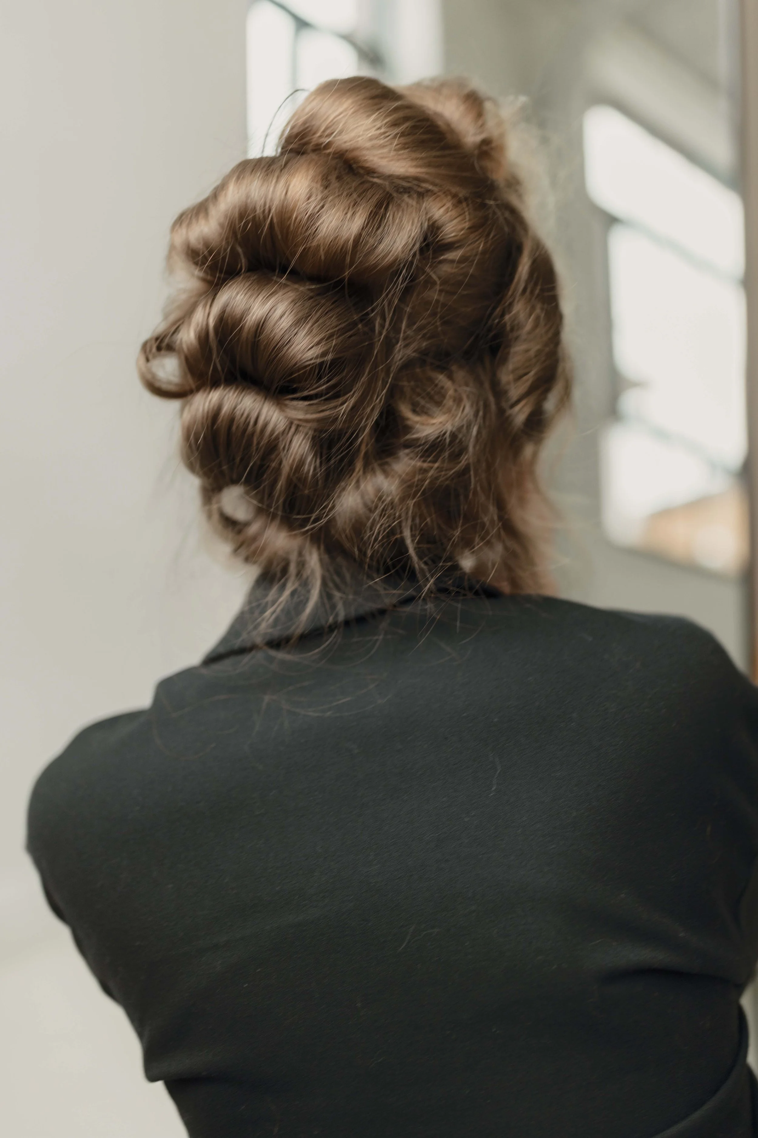 Back of a woman with light brown hair styled in an elegant updo, wearing a black top, standing near a window with natural light.