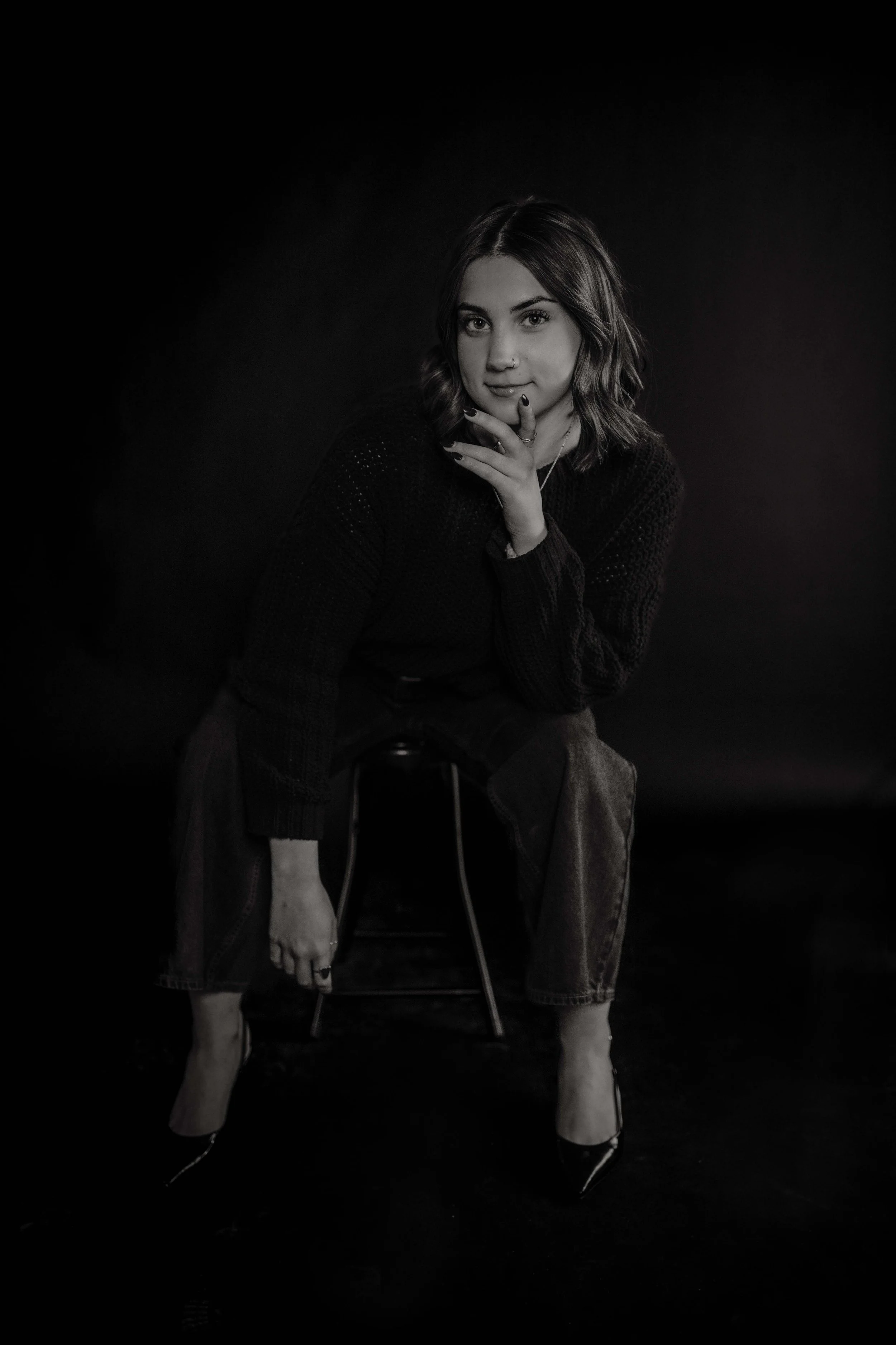 A young woman with wavy hair and a nose piercing, sitting on a stool, wearing a dark sweater, jeans, and high heels, with her hand touching her chin, in a black and white studio portrait.