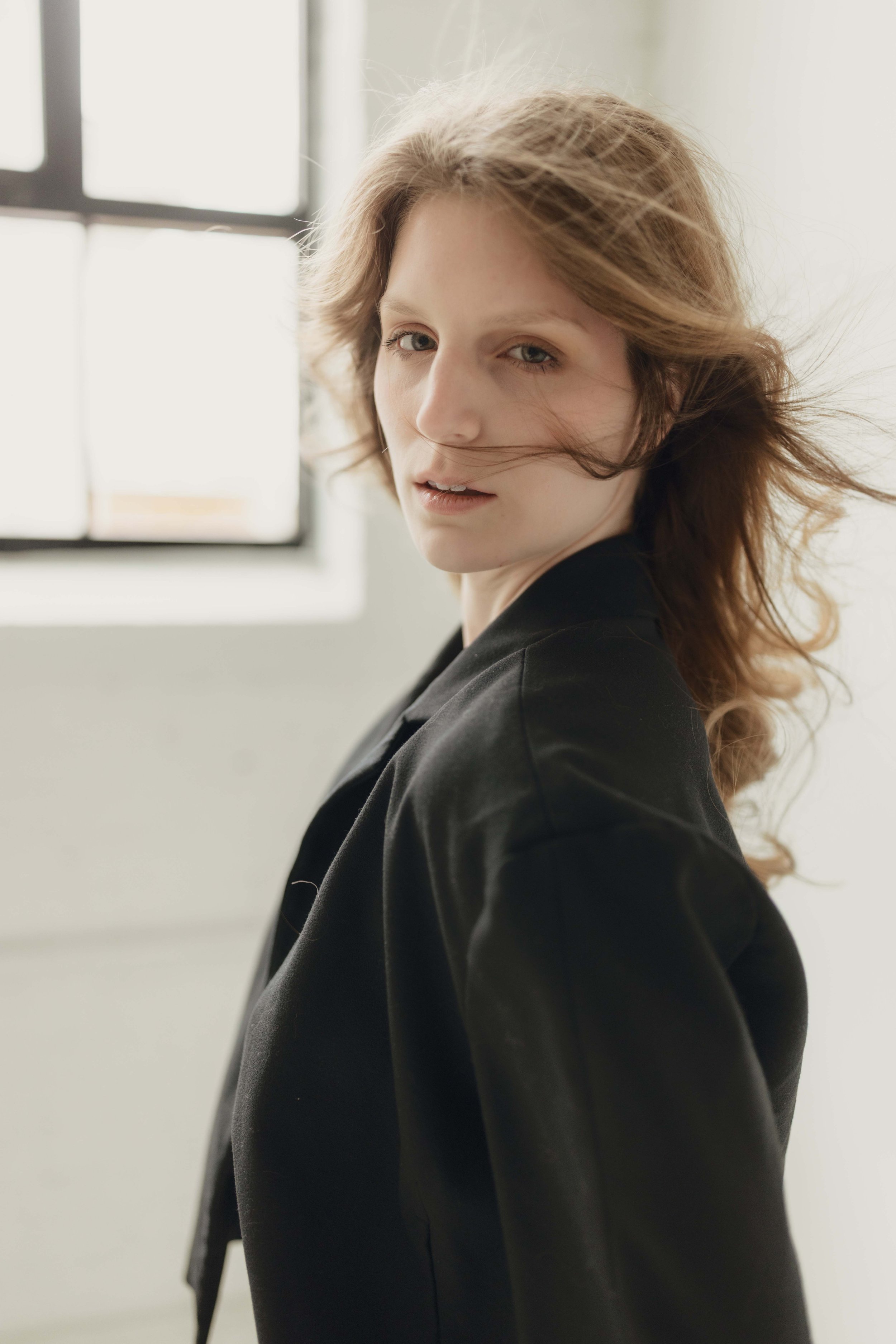 A woman with wavy, shoulder-length hair looking over her shoulder in front of a window with natural light.