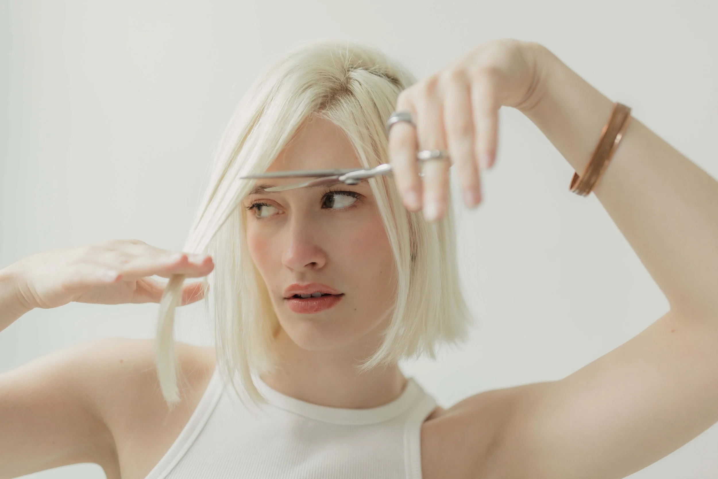 A blonde woman cutting her hair with scissors while looking in a mirror.