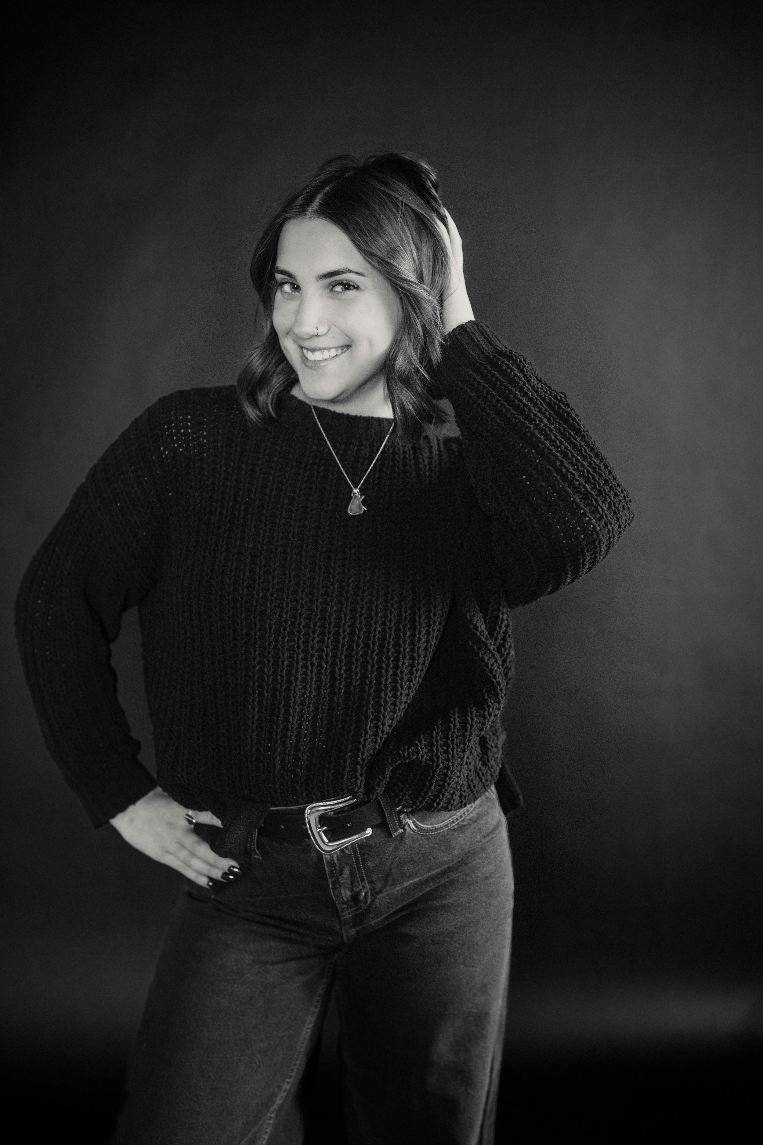 A young woman with shoulder-length hair, smiling, wearing a dark knitted sweater, necklace, and jeans, standing against a dark background, touching her hair.