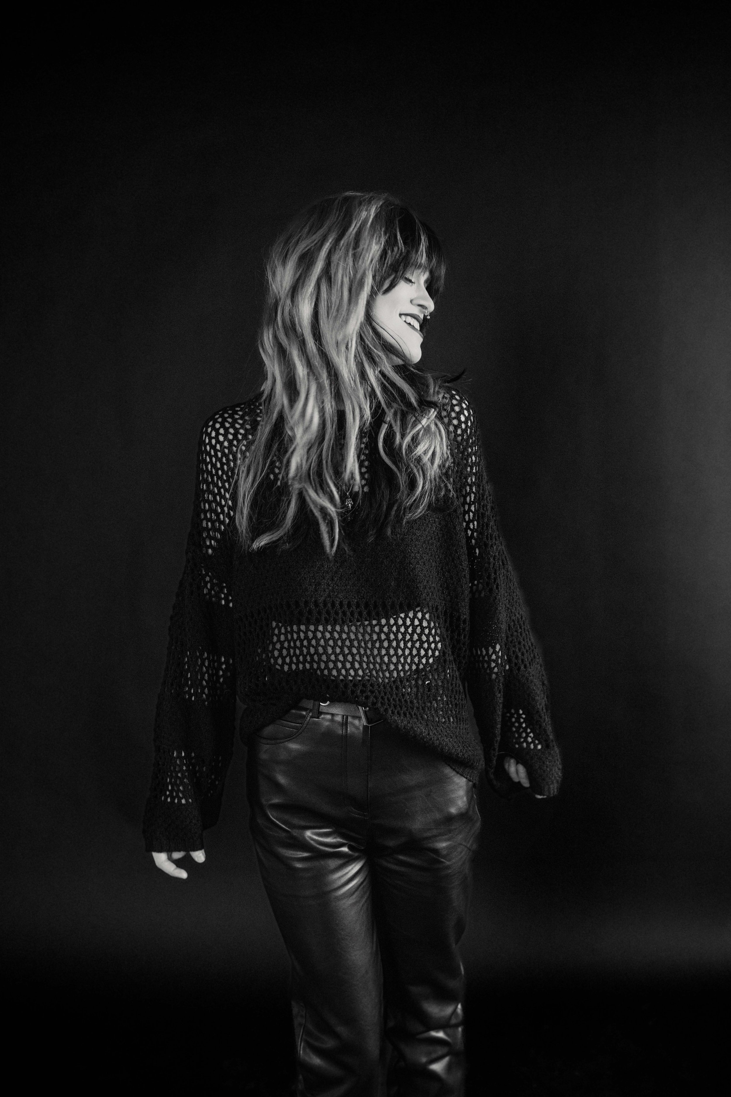 A woman with long wavy hair smiling and looking to the side, wearing a black crochet top and leather pants, standing against a dark background.