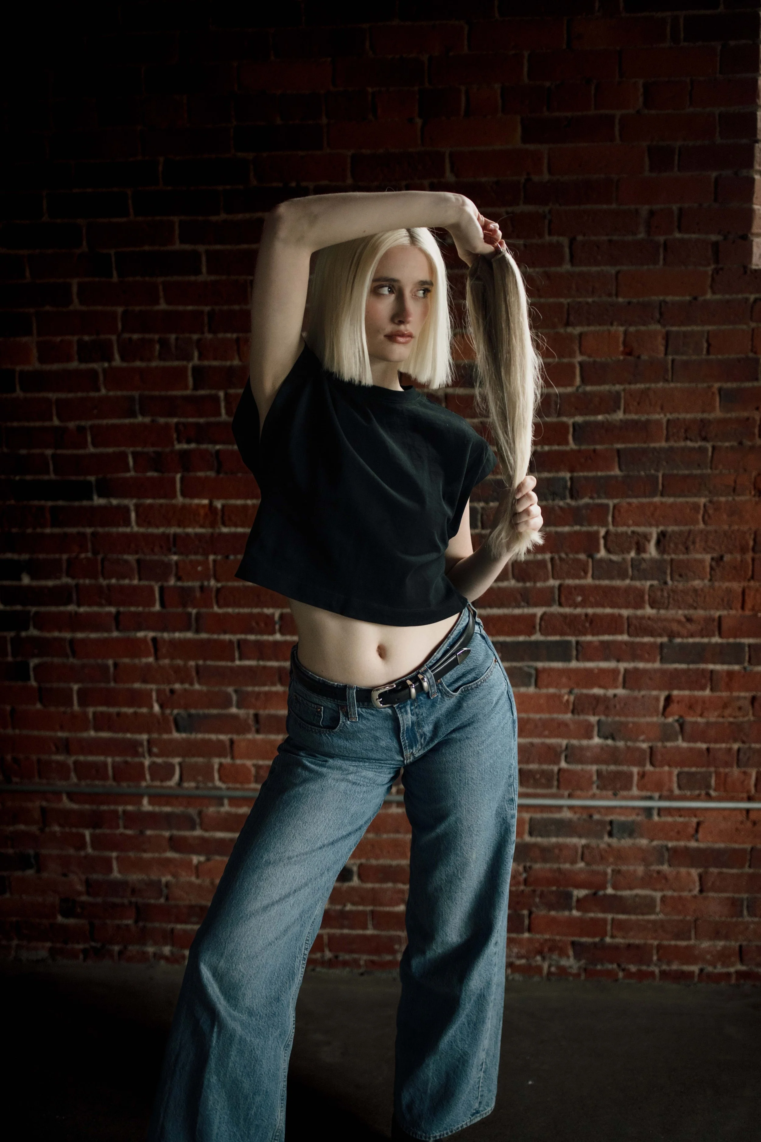 A woman with platinum blonde hair posing with one arm raised and holding her ponytail against a brick wall background, wearing a black crop top and loose blue jeans.