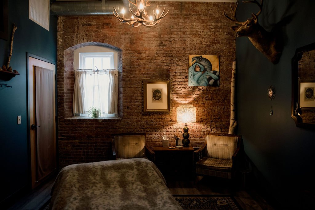 A cozy bedroom with exposed brick walls, an arched window with curtains, a chandelier, two armchairs beside a side table with a lamp, framed artwork, a mounted deer head, and a mirror on the wall.
