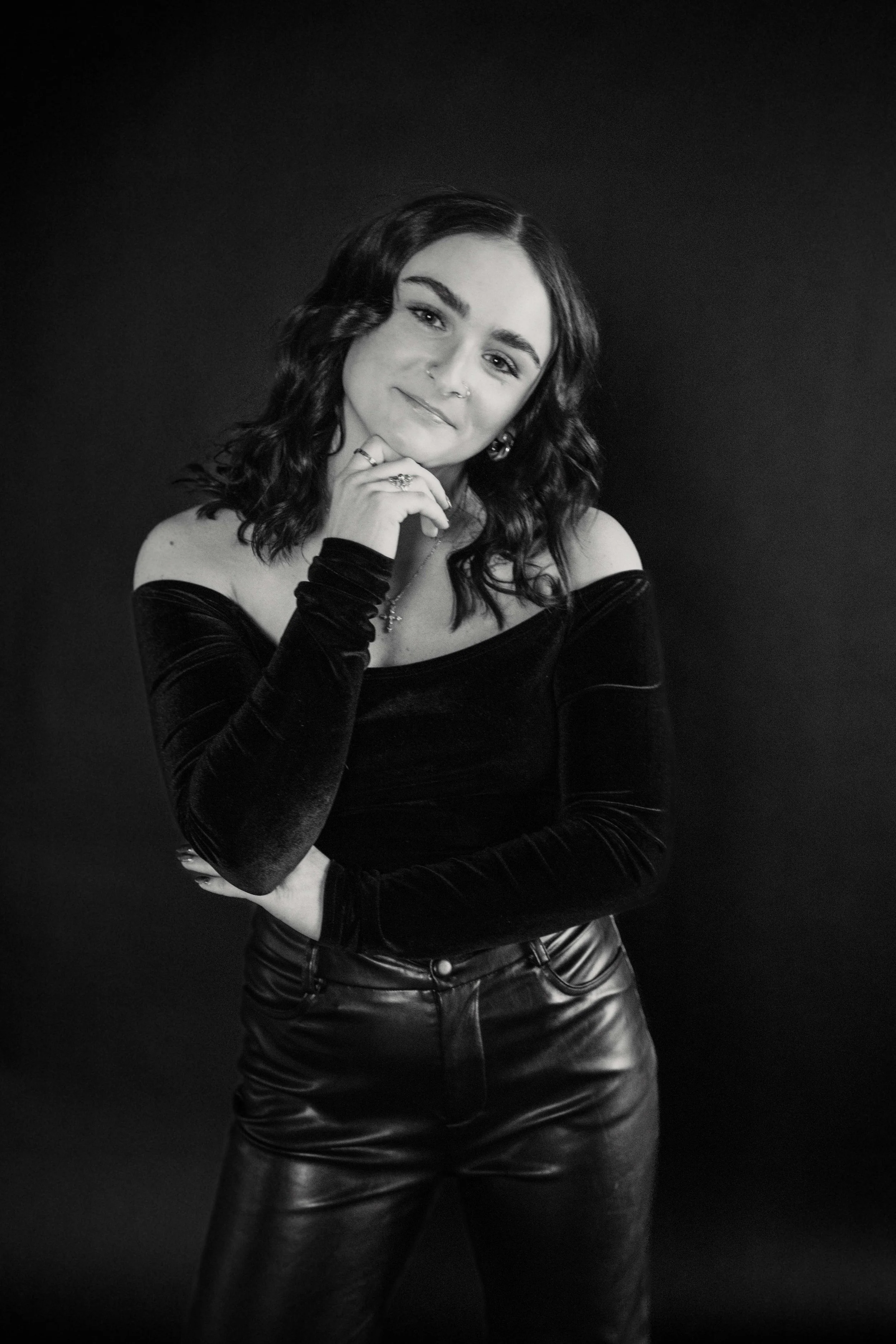 A young woman with wavy dark hair, wearing a off-the-shoulder black top and leather pants, standing against a plain dark background, looking at the camera with a slight smile.