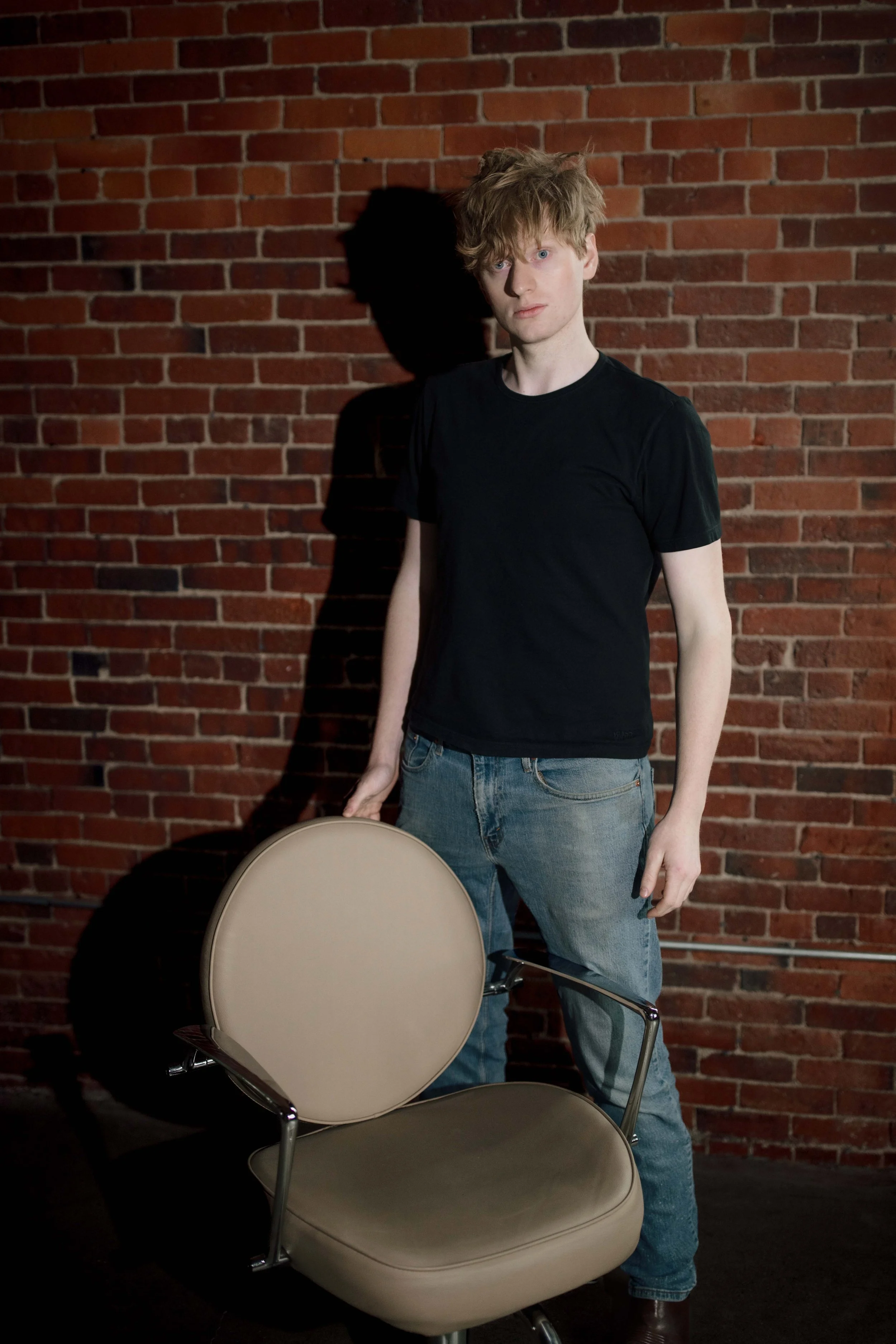 A young man with curly, tousled brown hair and fair skin standing in front of a red brick wall. He is wearing a black t-shirt and blue jeans, holding a beige chair with metal legs. Part of a black tire and another chair are visible in the foreground.