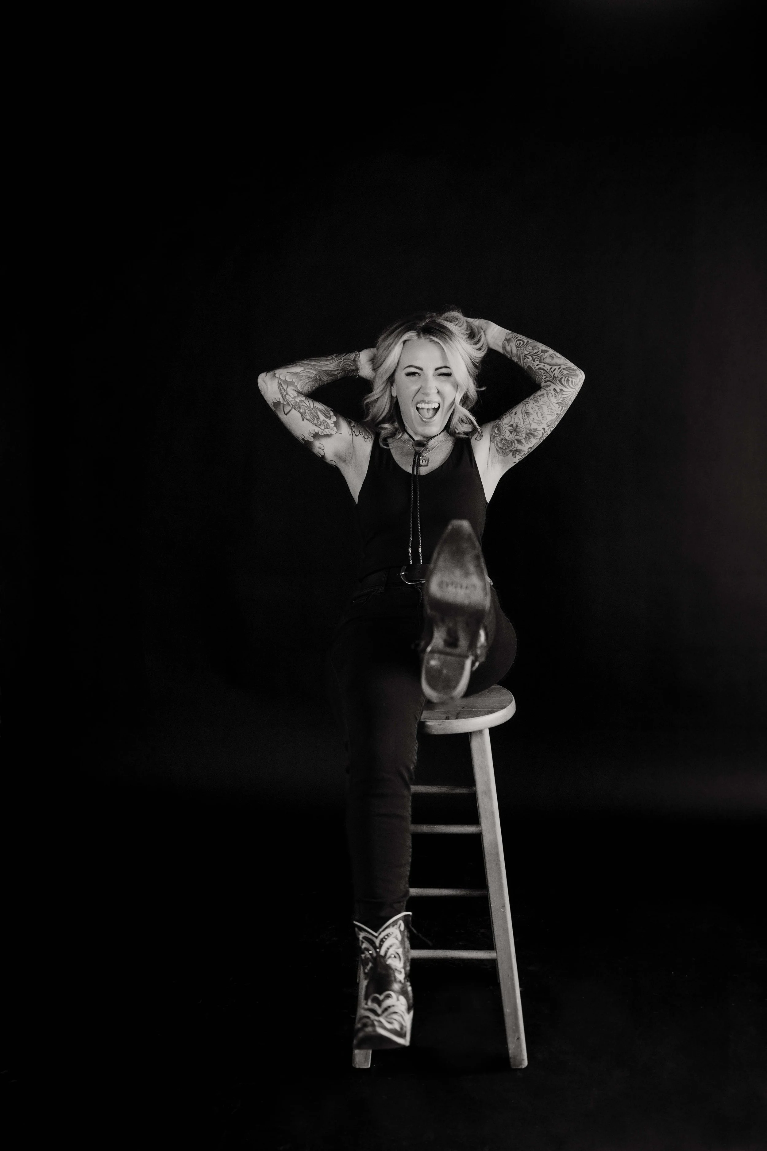 Black and white photo of a woman with tattooed arms, holding her hair and sticking out her tongue, sitting on a stool with one leg crossed over the other, wearing a tank top and sneakers, with a dark background.