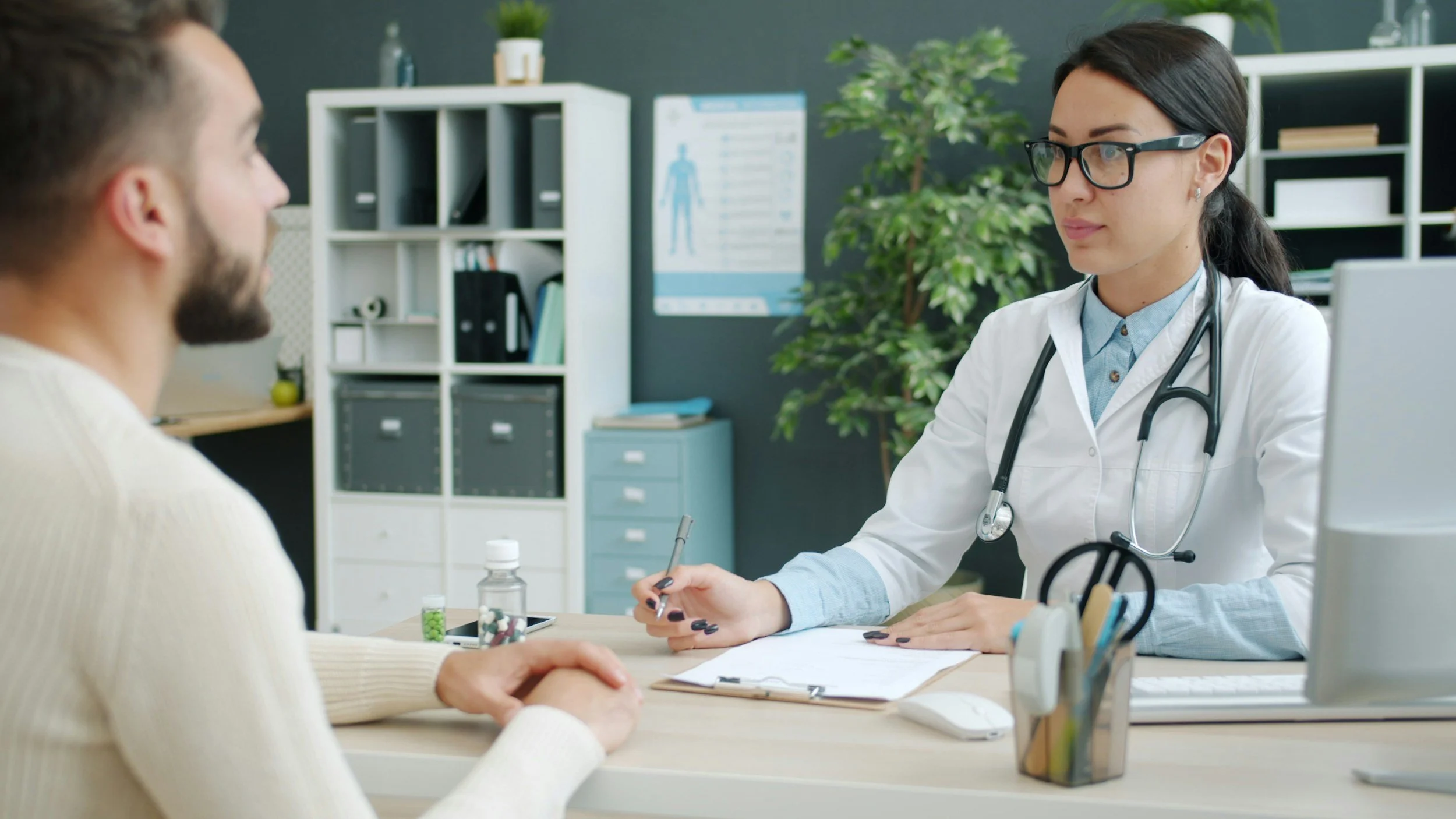 A doctor in a white coat and glasses speaking with a patient in an office with shelves, a plant, and medical posters in the background.