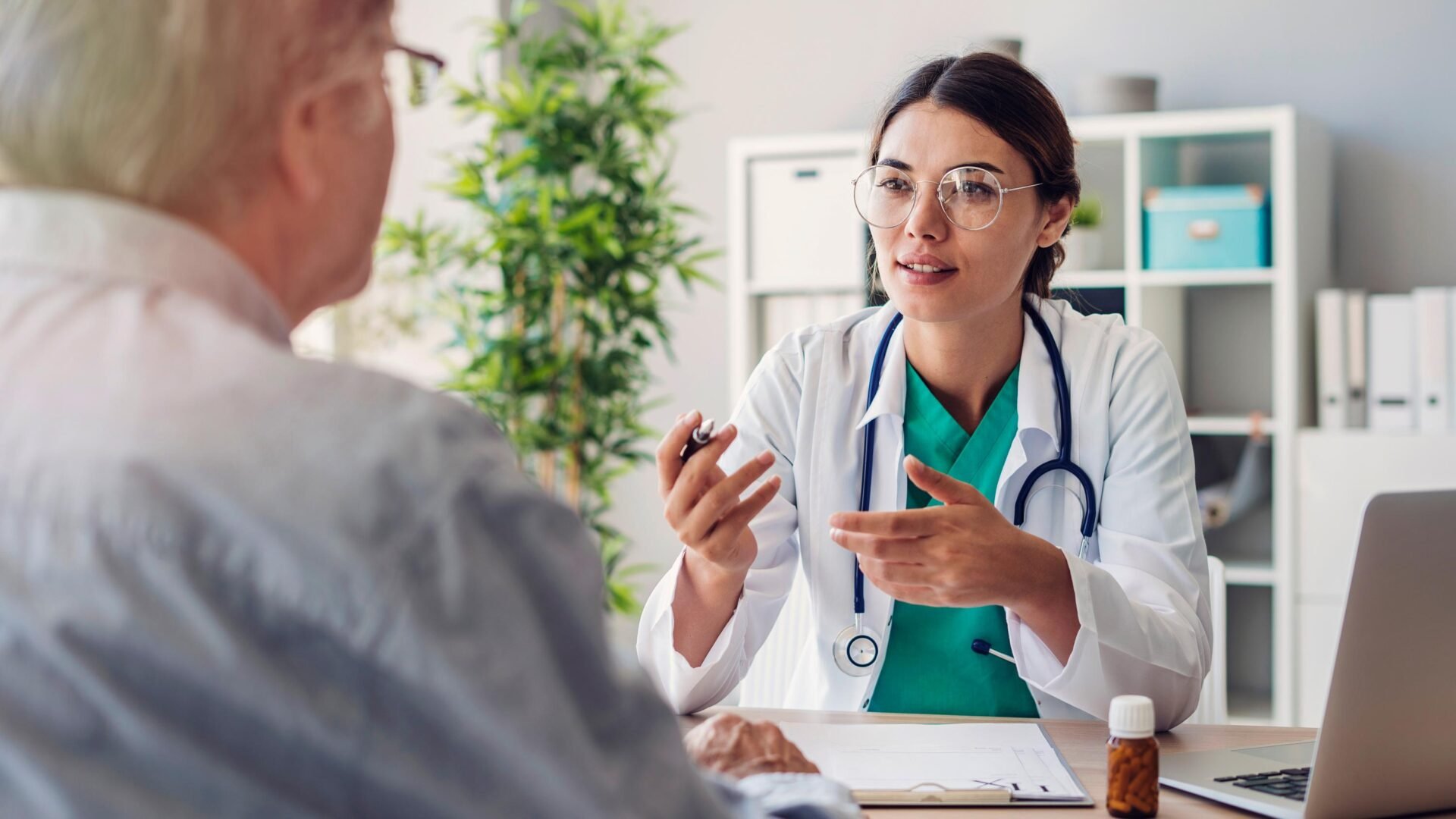 Medical professional and a patient in consultation. The doctor appears to be explaining or discussing, with medical supplies and a computer in the background.