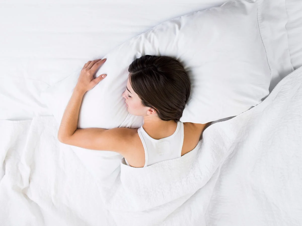 A woman with short dark hair sleeping on her side in a white bed, resting her head on a white pillow.