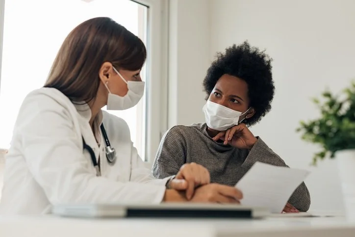 A doctor and a patient, both wearing masks, sitting at a table in a medical office. The doctor is holding a clipboard or paper, and they appear to be having a conversation.
