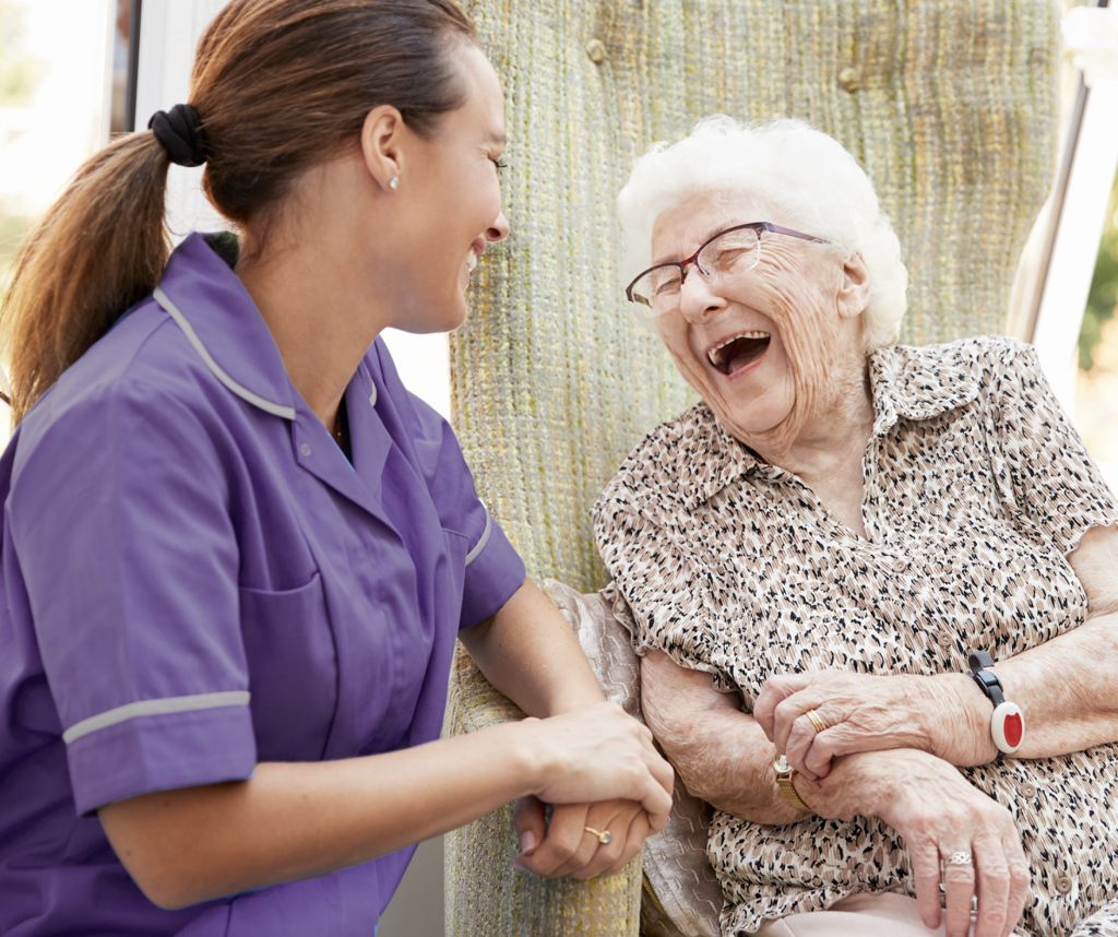 A caregiver woman in purple uniform talking and laughing with an elderly woman with glasses, wearing a leopard print shirt, who is also laughing.
