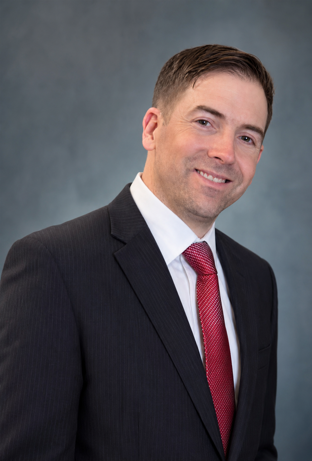 A man wearing a dark business suit with pinstripes, white shirt, and red tie, smiling against a gray backdrop.
