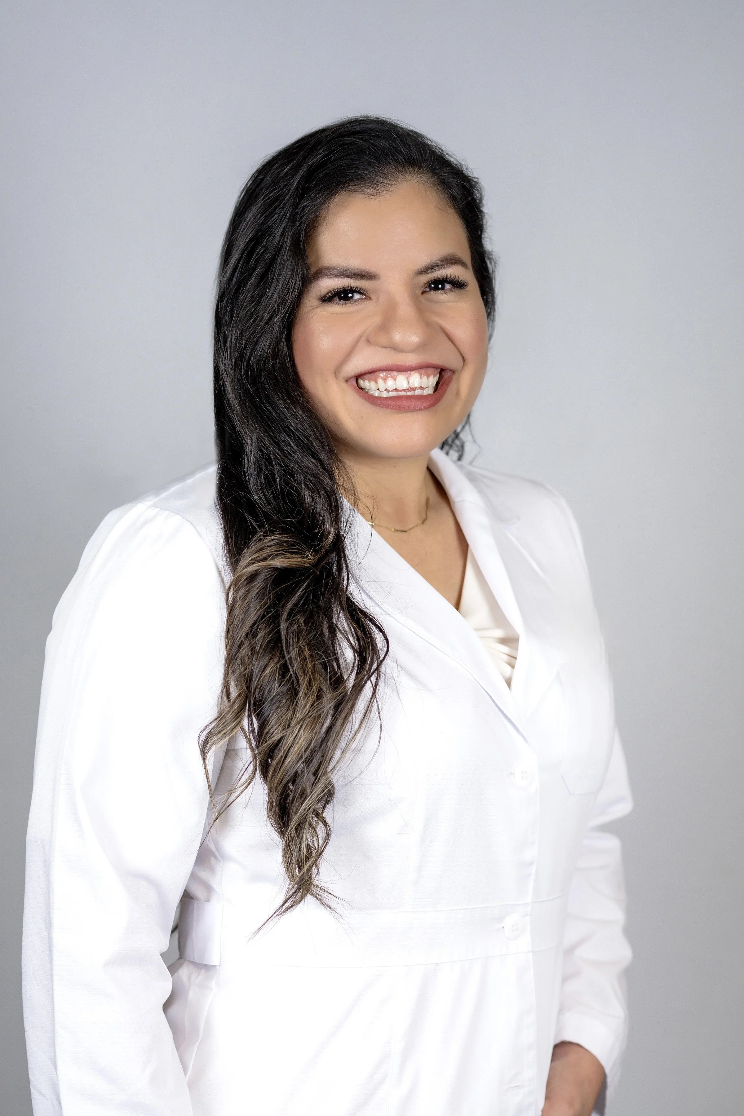 A smiling woman with long dark wavy hair wearing a white coat, standing against a gray background.