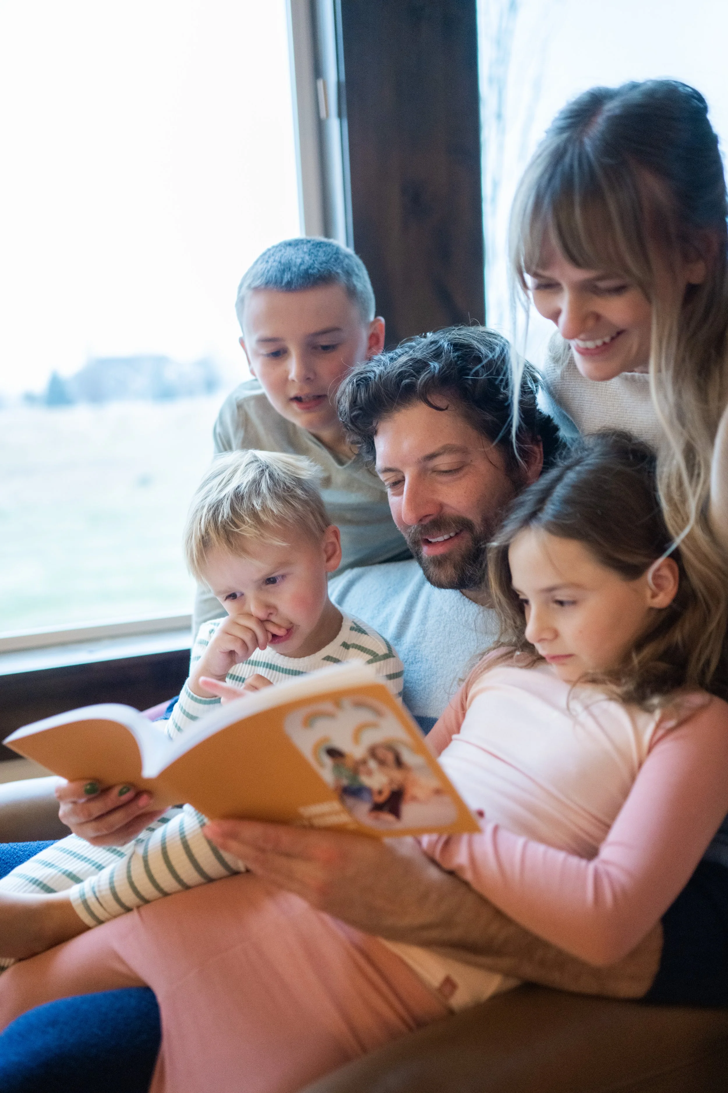 A family of five people, including three children, reading a Jambook together on a couch near a window in a cozy living room.