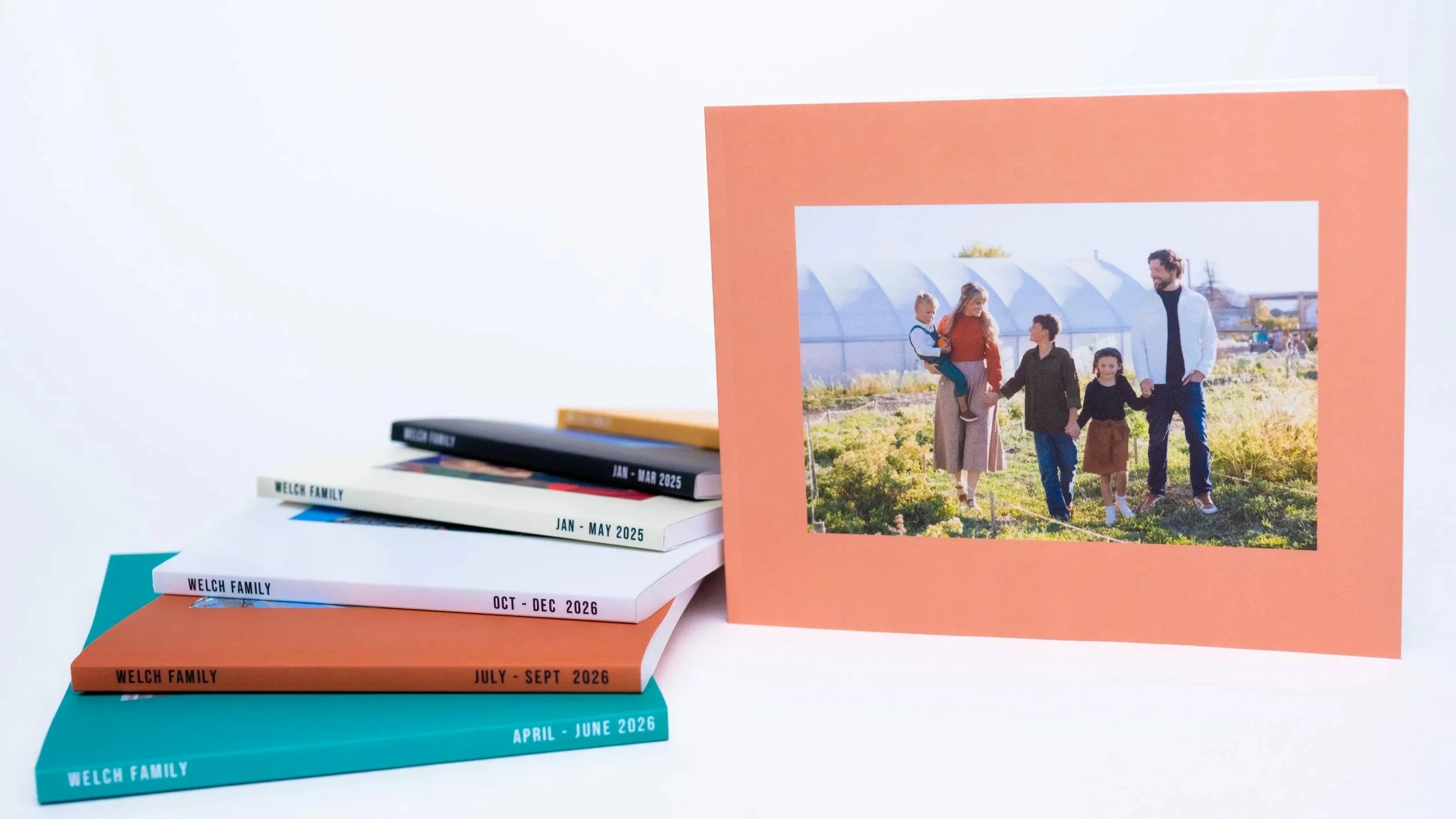 A family photo printed on an orange photo book, showing four people walking outside in a garden or farm setting. The image is displayed on top of a stack of photo books labeled with months and years.