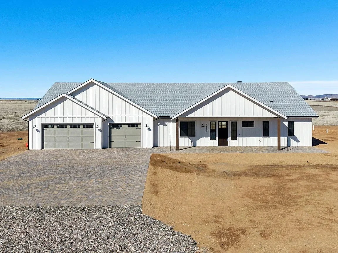Front view of a newly constructed modern house with white siding, grey garage doors, a small porch, and a paved driveway, set against a clear blue sky and a dry open landscape.