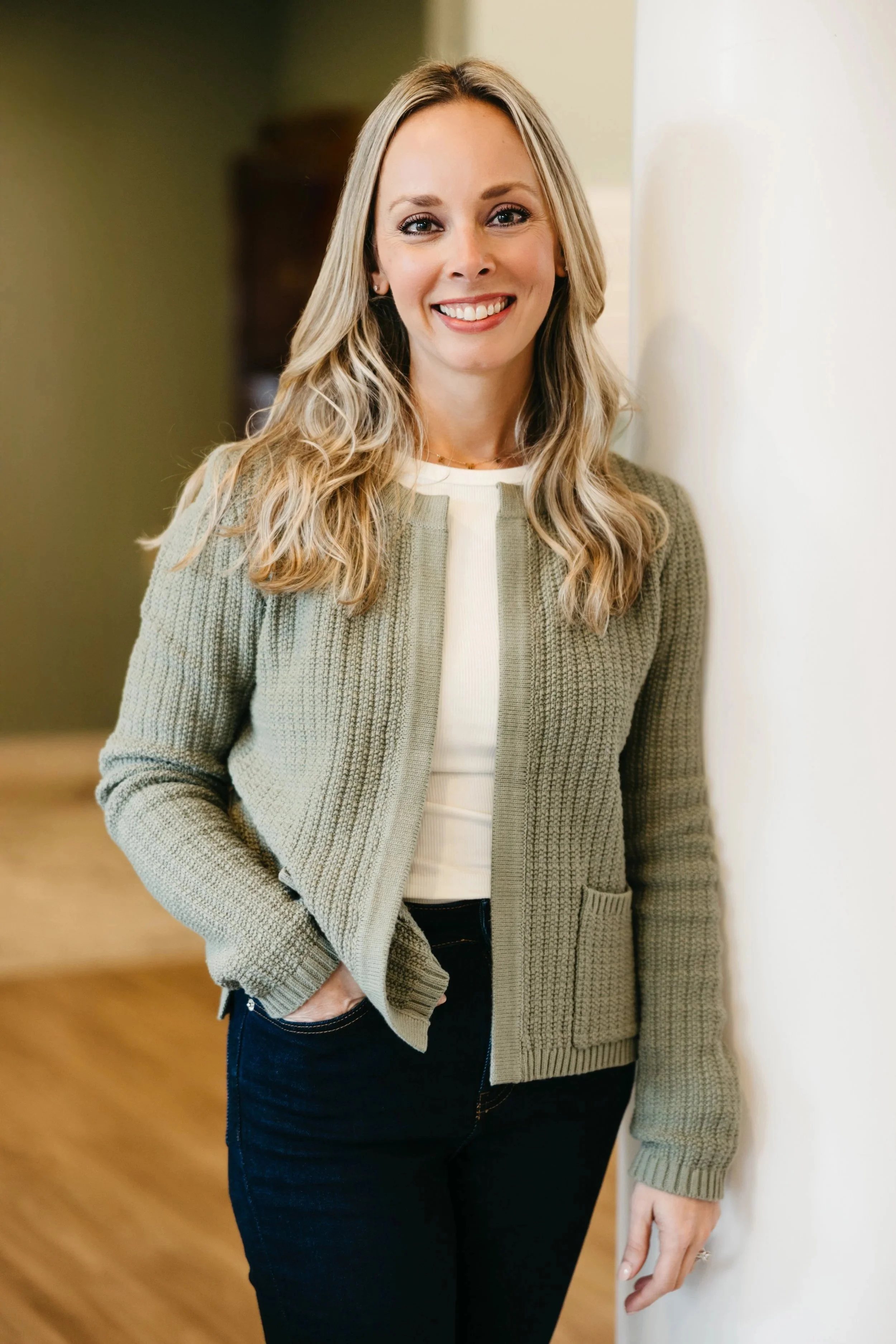 A woman with long blonde hair, smiling, wearing a beige knit sweater over a white top, dark jeans, standing indoors against a light-colored wall.