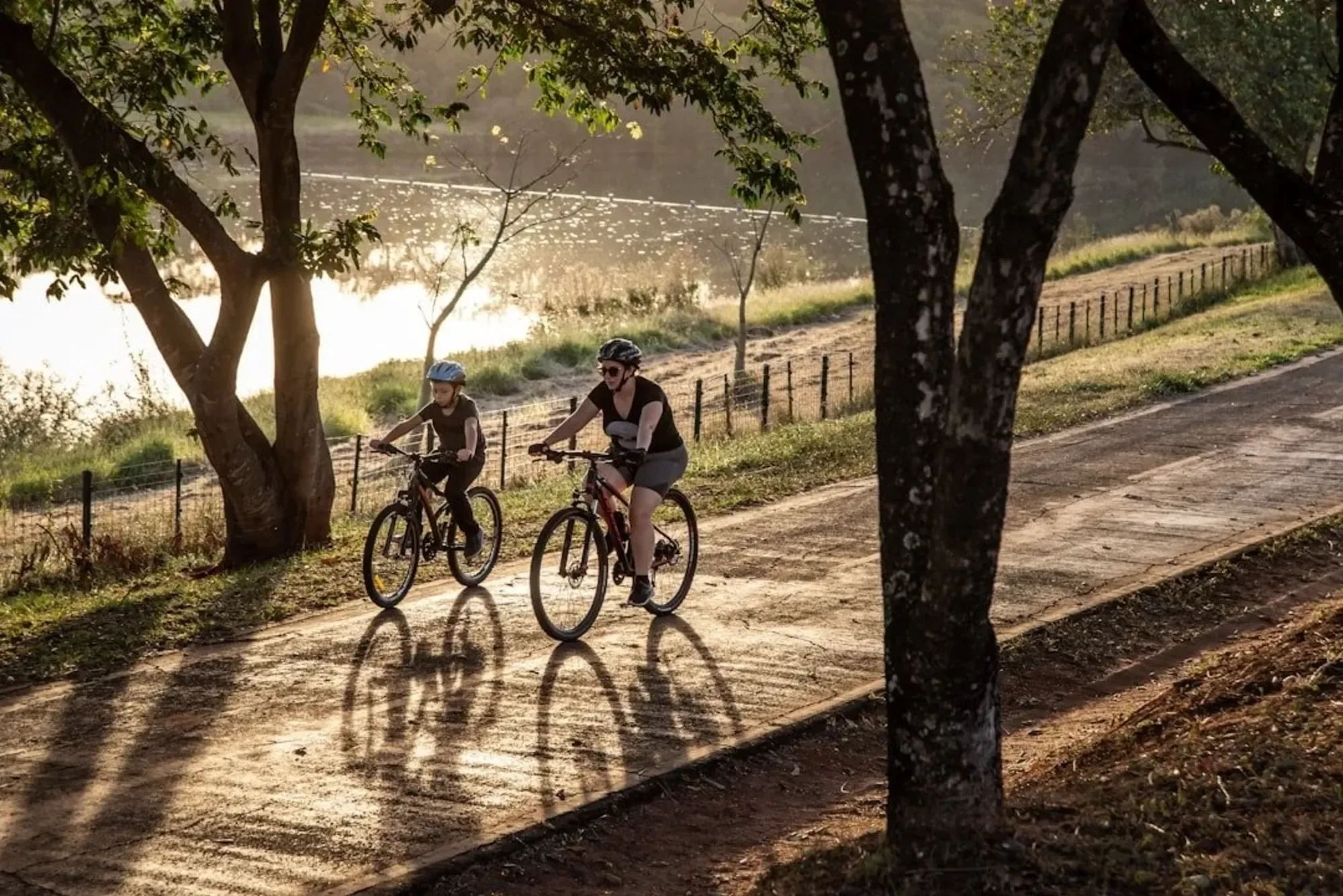 Two women and a child riding bicycles along a dirt path next to a body of water, with trees and a fence nearby in a park setting during late afternoon or early evening.