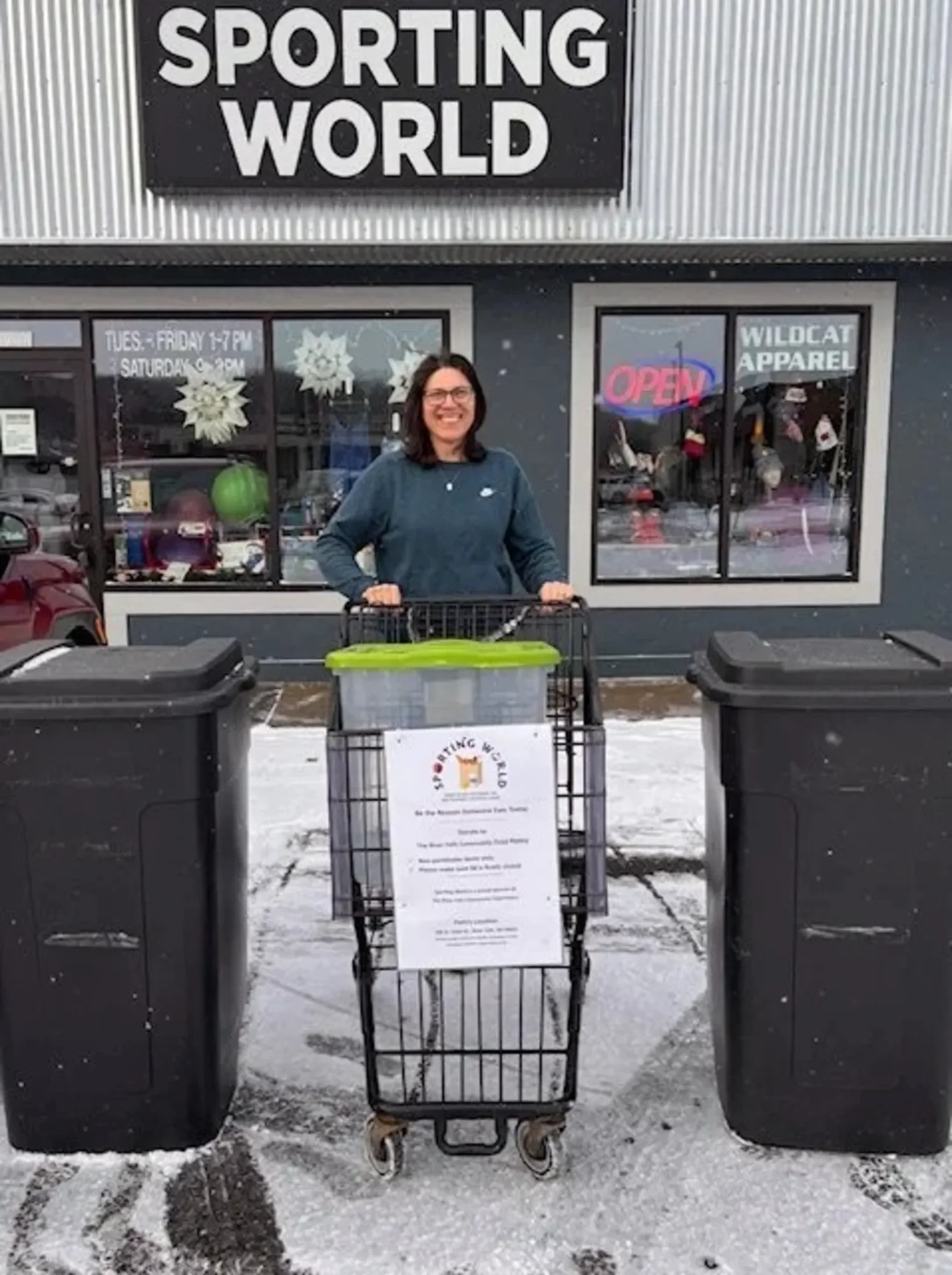 A smiling woman standing behind a shopping cart outside a store called 'Sporting World'. The store has large windows with an 'Open' sign in pink and white, and snow on the ground.