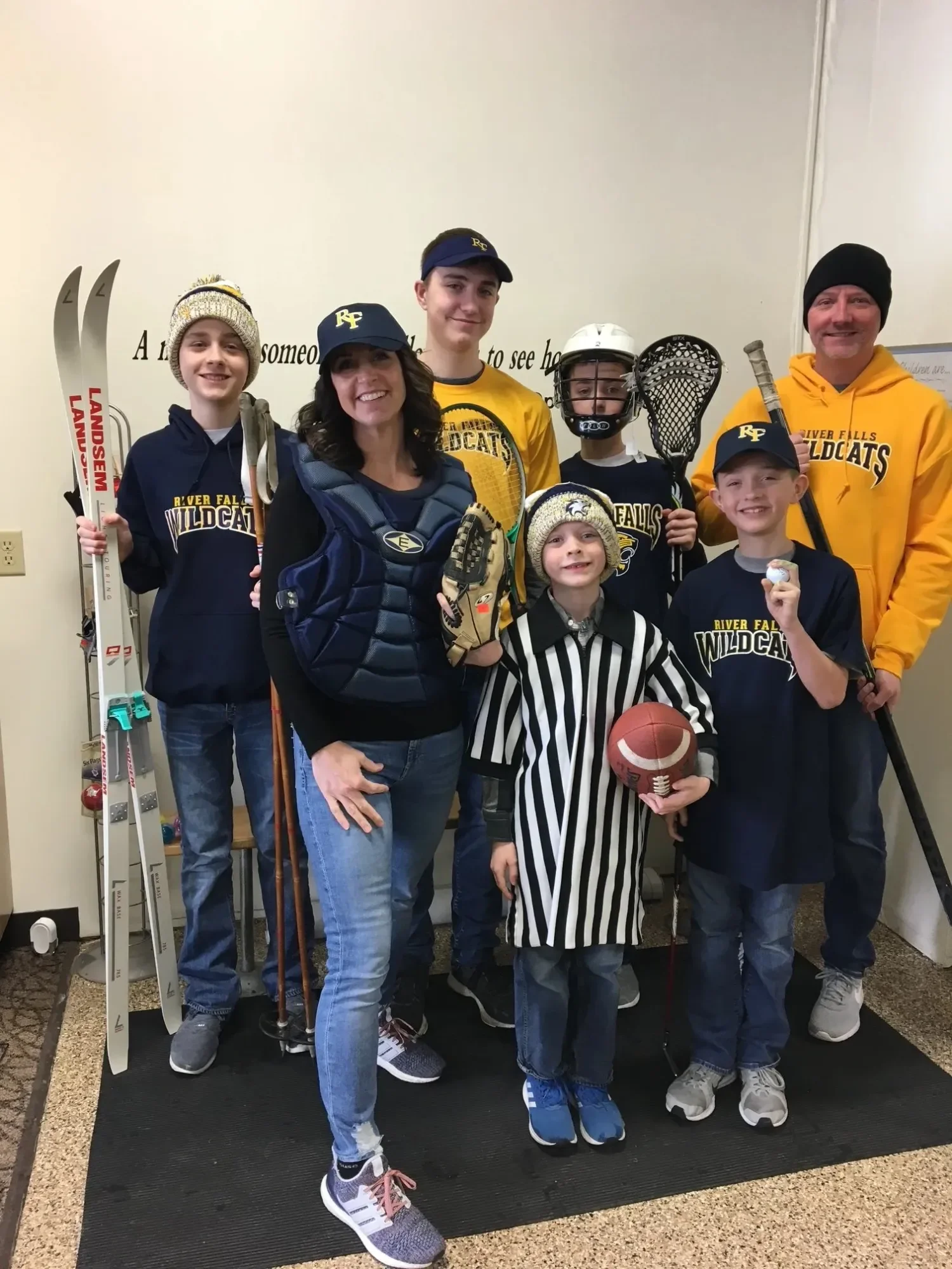 Group of seven people, including children and adults, dressed in sports gear and hockey apparel, standing in front of a wall with words on it, holding hockey sticks, a football, and wearing hockey helmets and hats.
