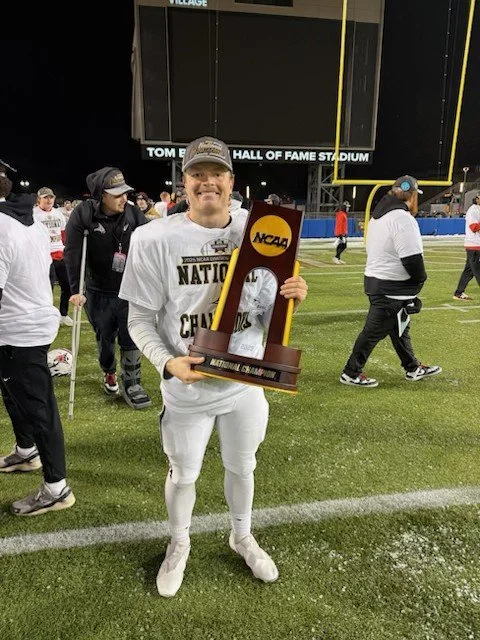 A smiling baseball player in uniform holding an NCAA national championship trophy on a stadium field surrounded by people celebrating.